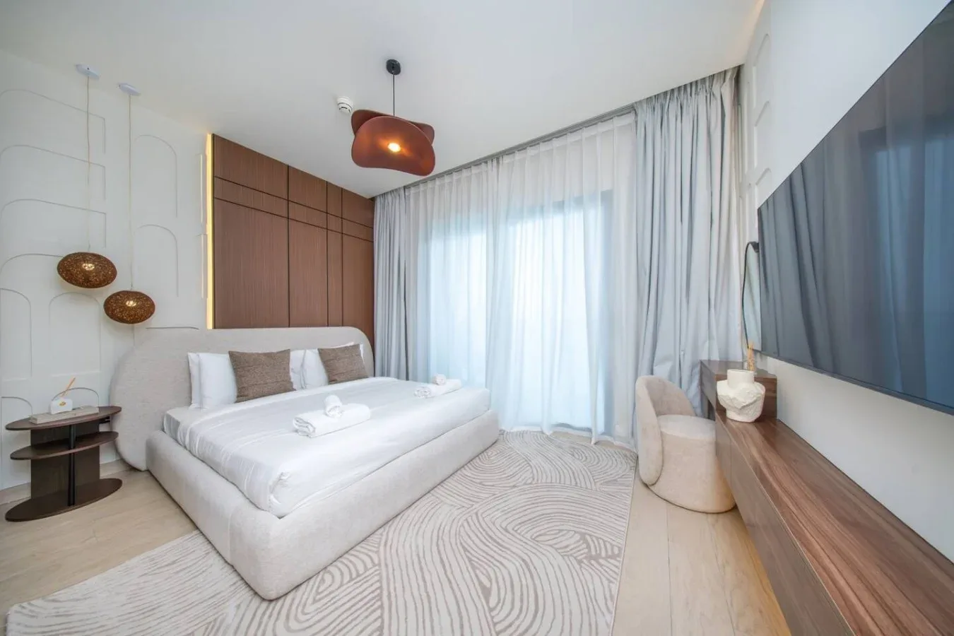 Un dormitorio luminoso tiene una cama junto a cortinas translúcidas y un televisor.
