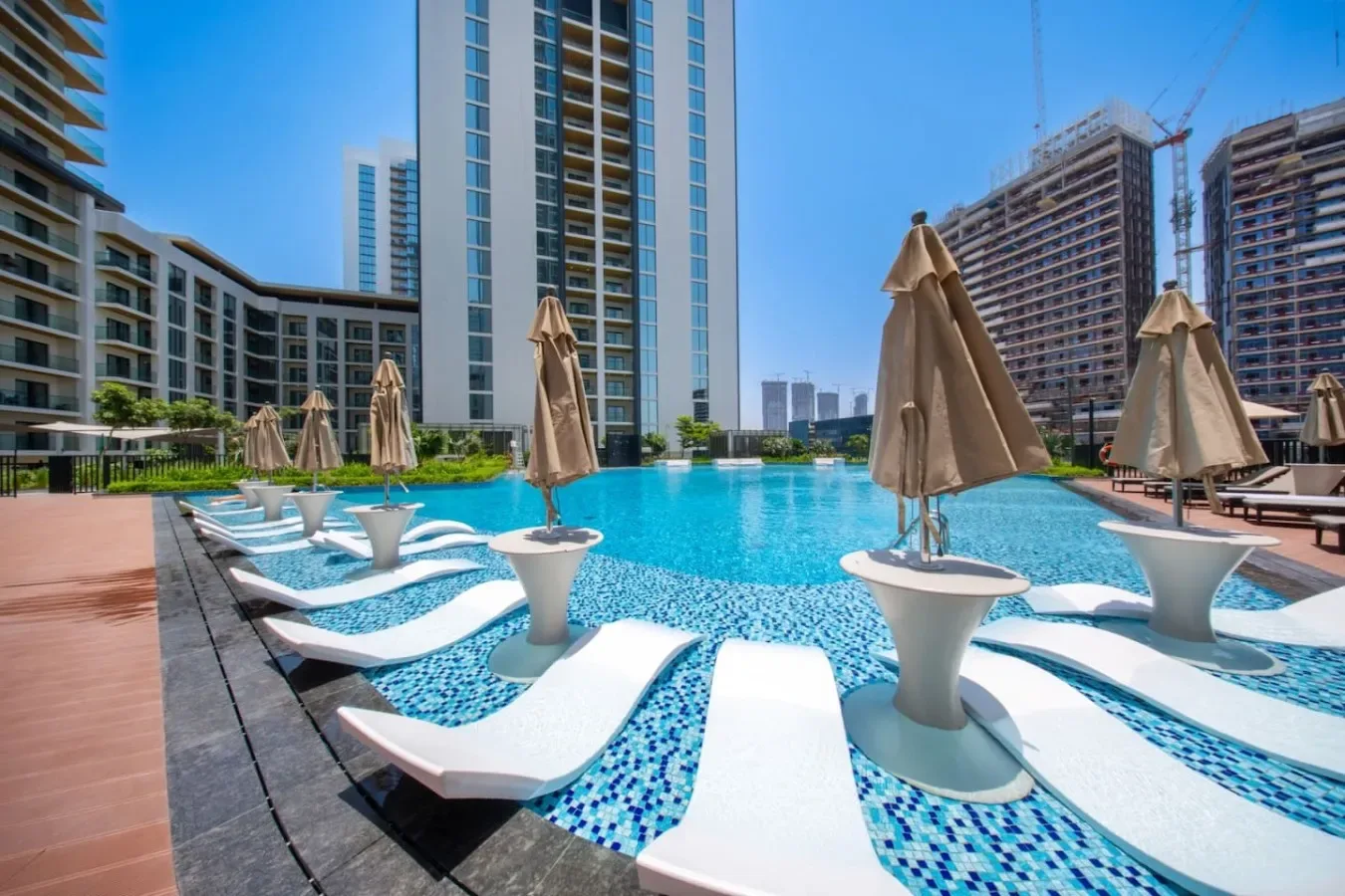 Una piscina con tumbonas blancas curvas y sombrillas cerradas está rodeada de edificios altos.