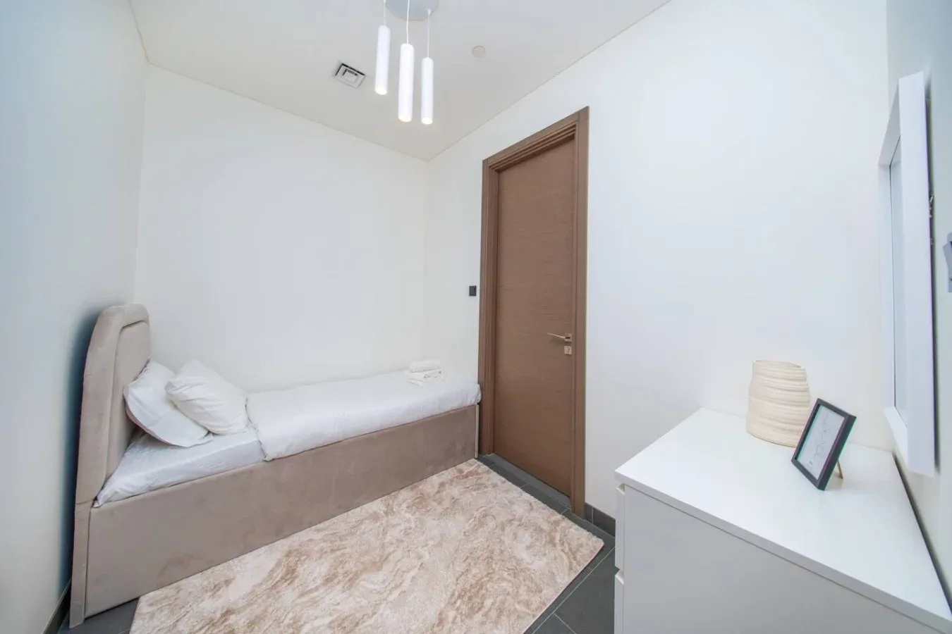 Un dormitorio luminoso con una cama individual, una puerta de madera cerrada y una cómoda blanca.