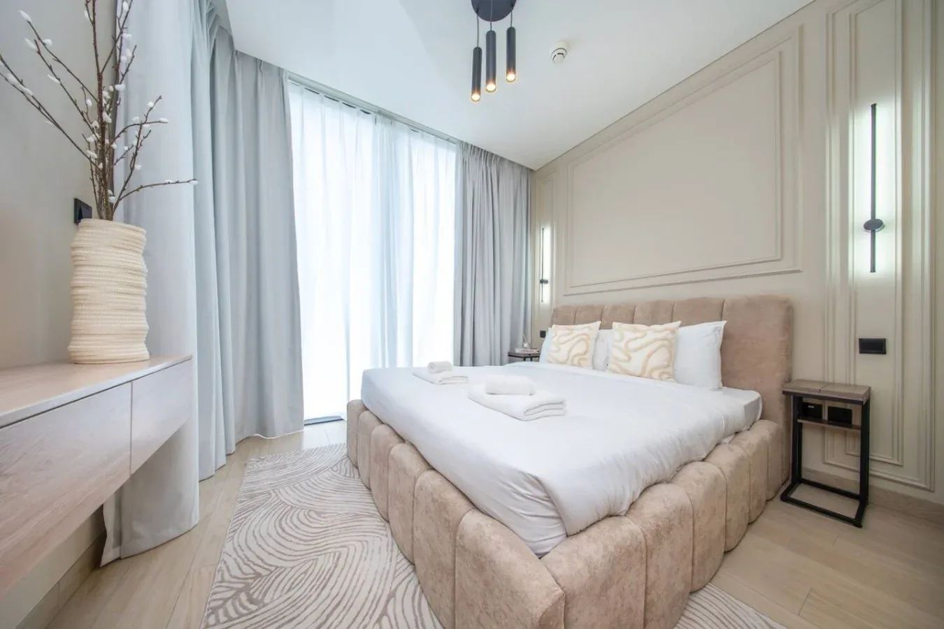 Un dormitorio luminoso con una cama grande, cortinas claras y una alfombra estampada.