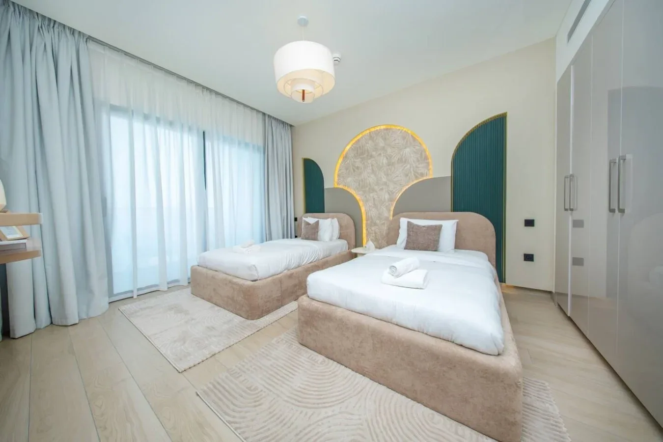 Un dormitorio luminoso con dos camas, cortinas translúcidas y un detalle decorativo en la pared.