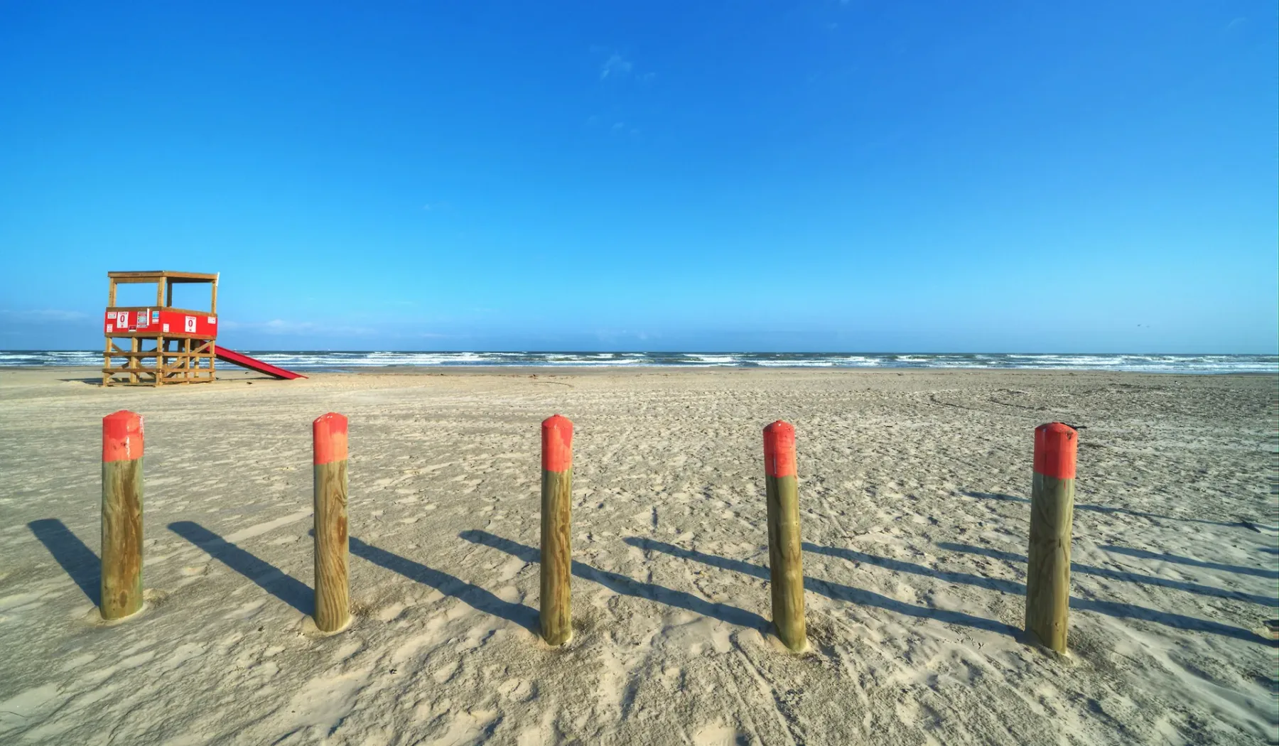 A sandy beach with a red lifeguard tower and posts in the foreground.