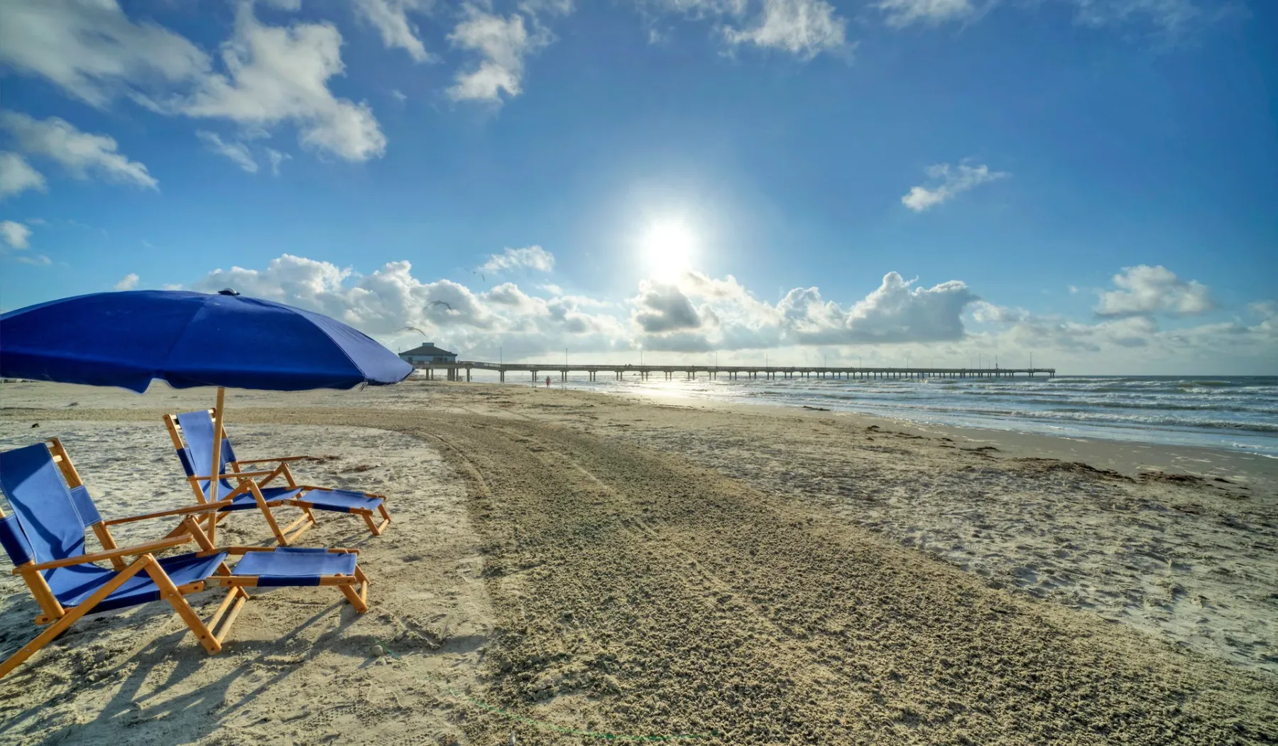 A sunny beach with lounge chairs beneath a blue umbrella and the ocean beyond.