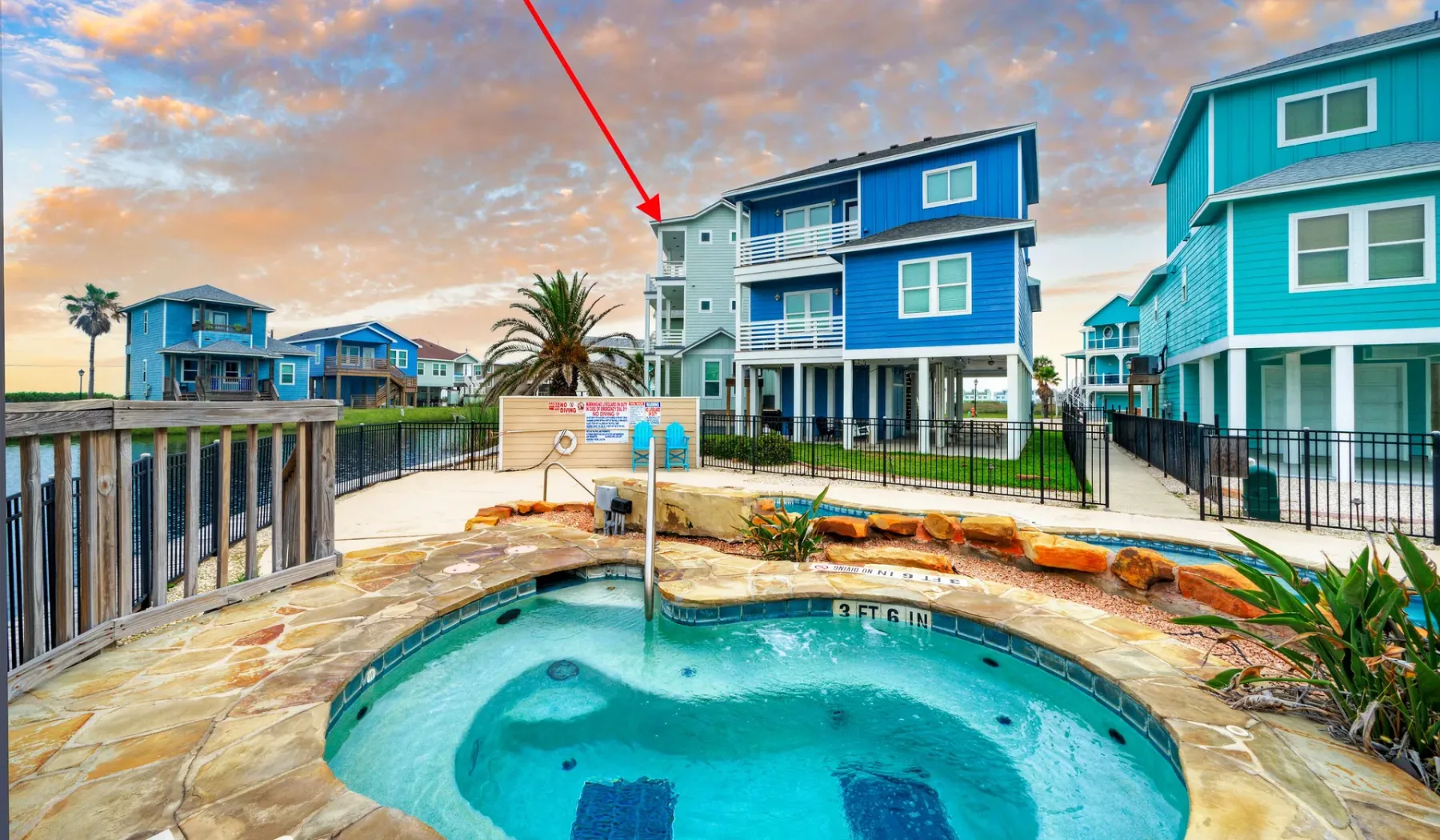 An outdoor swimming pool sits beside colorful coastal-style houses under a cloudy sky.