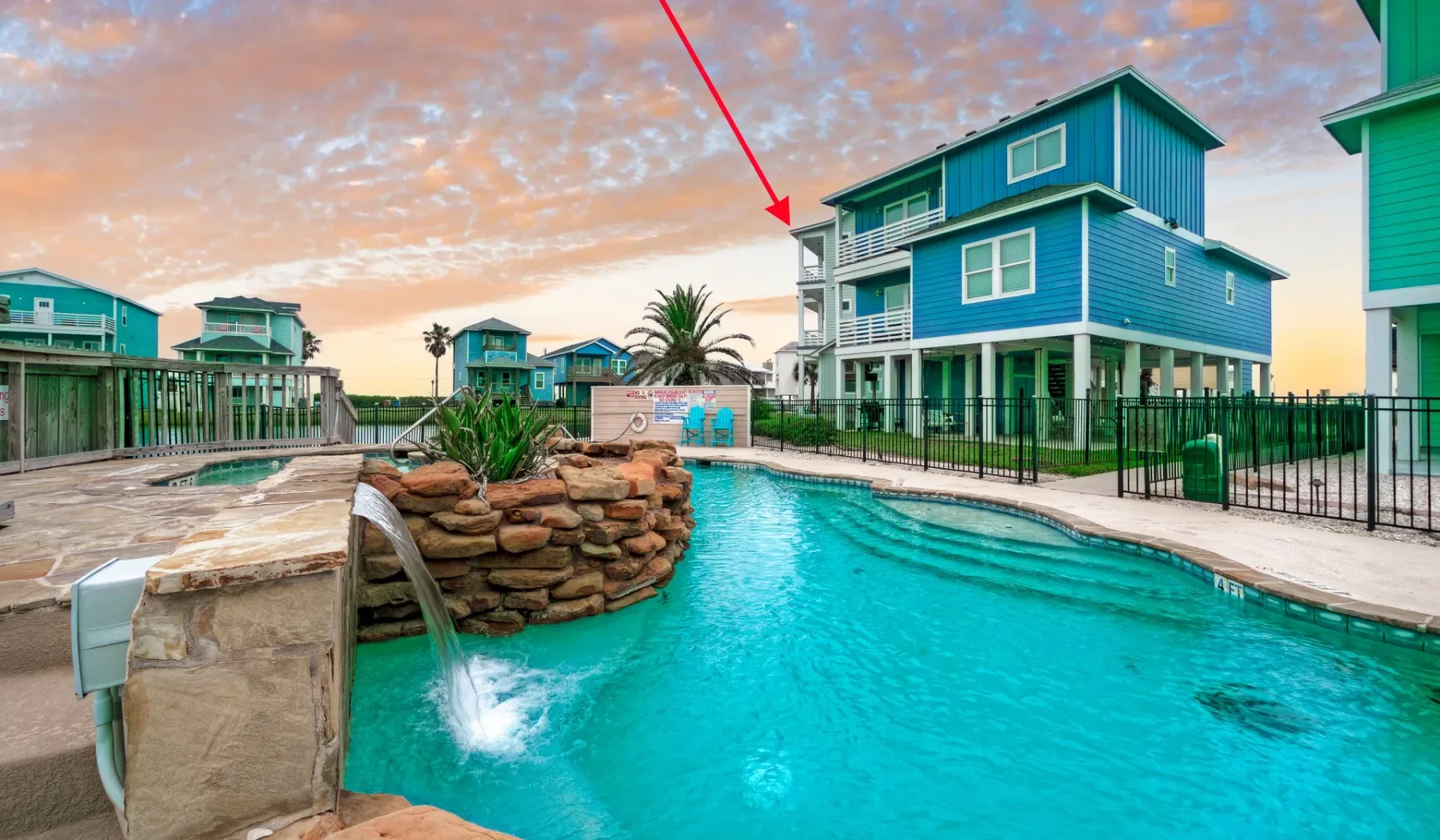 A swimming pool curves around a rock feature beside a blue coastal-style building.