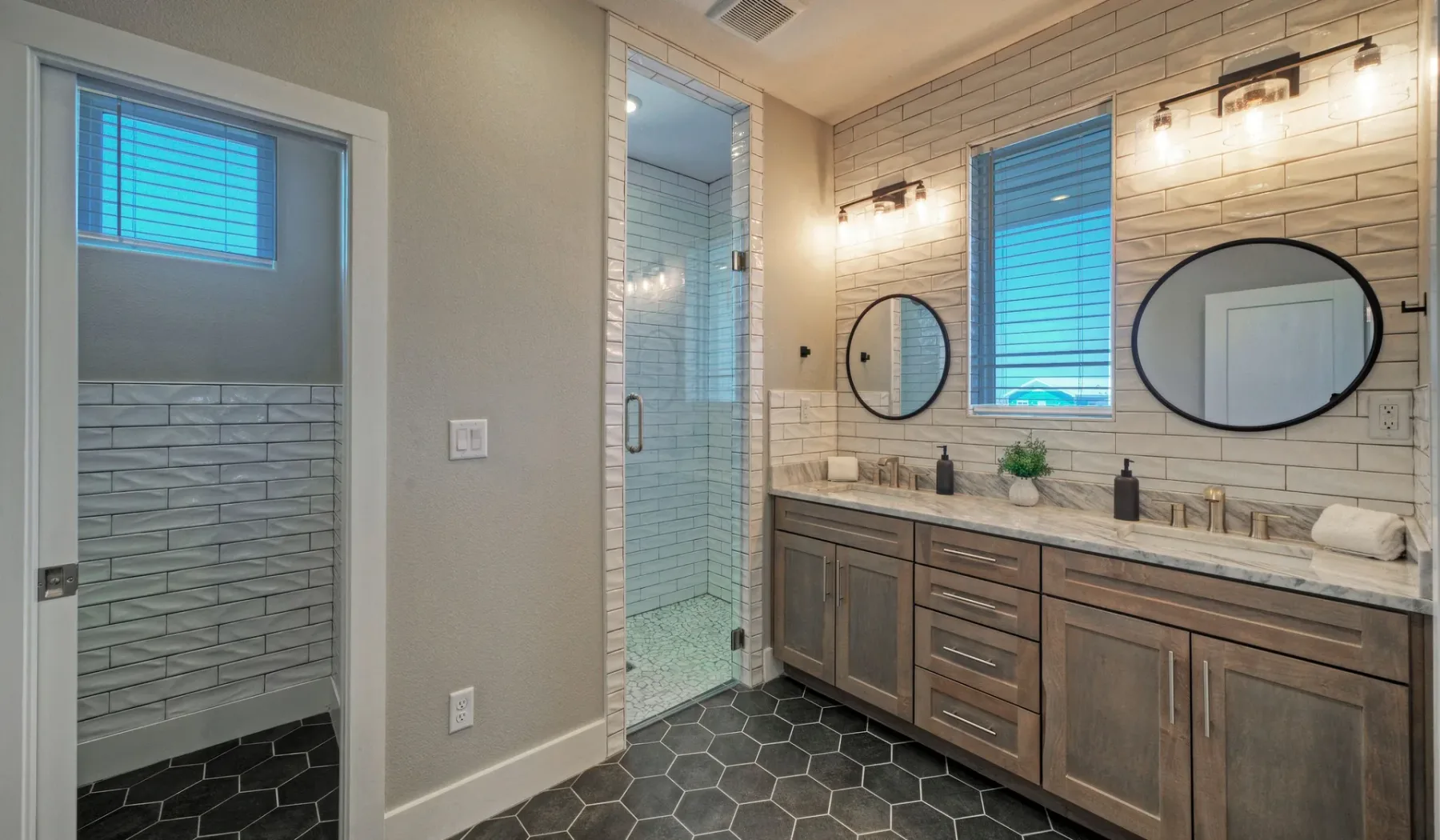 A bathroom vanity with two mirrors, a tiled shower, and dark hexagonal floor tiles.