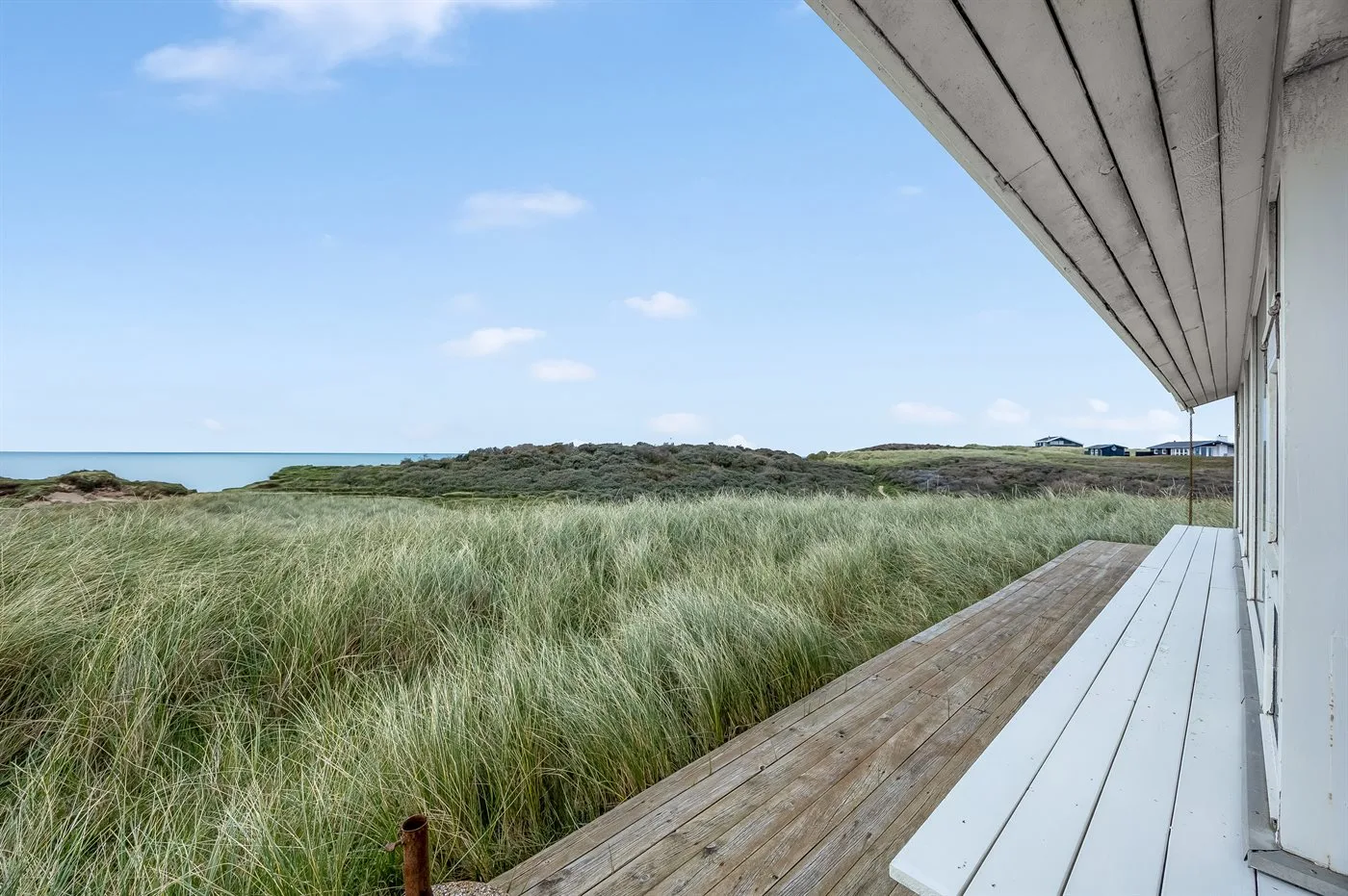 Una terraza de madera da a dunas cubiertas de hierba y al océano bajo un cielo azul.