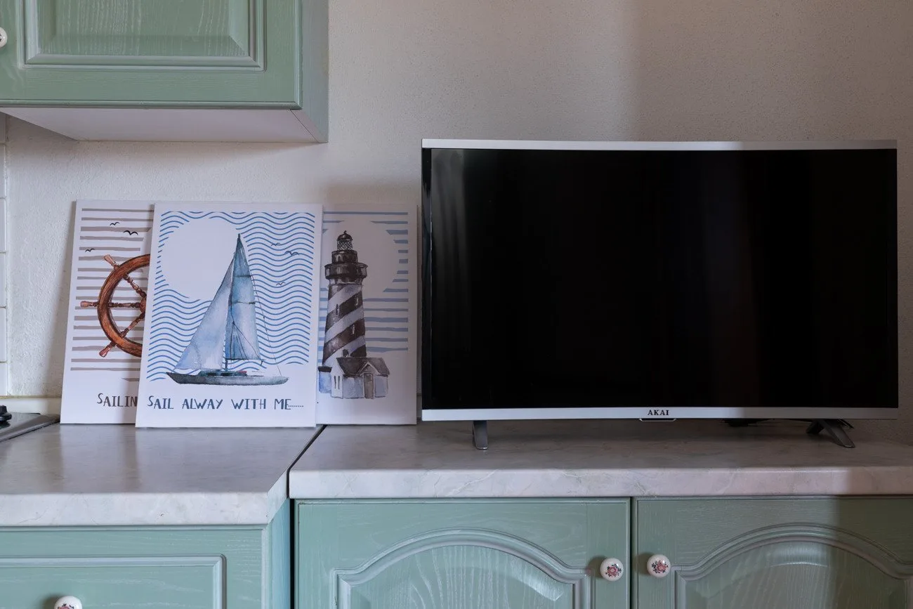 A television sits on a pale countertop above mint-green cabinets, with nautical prints beside it.