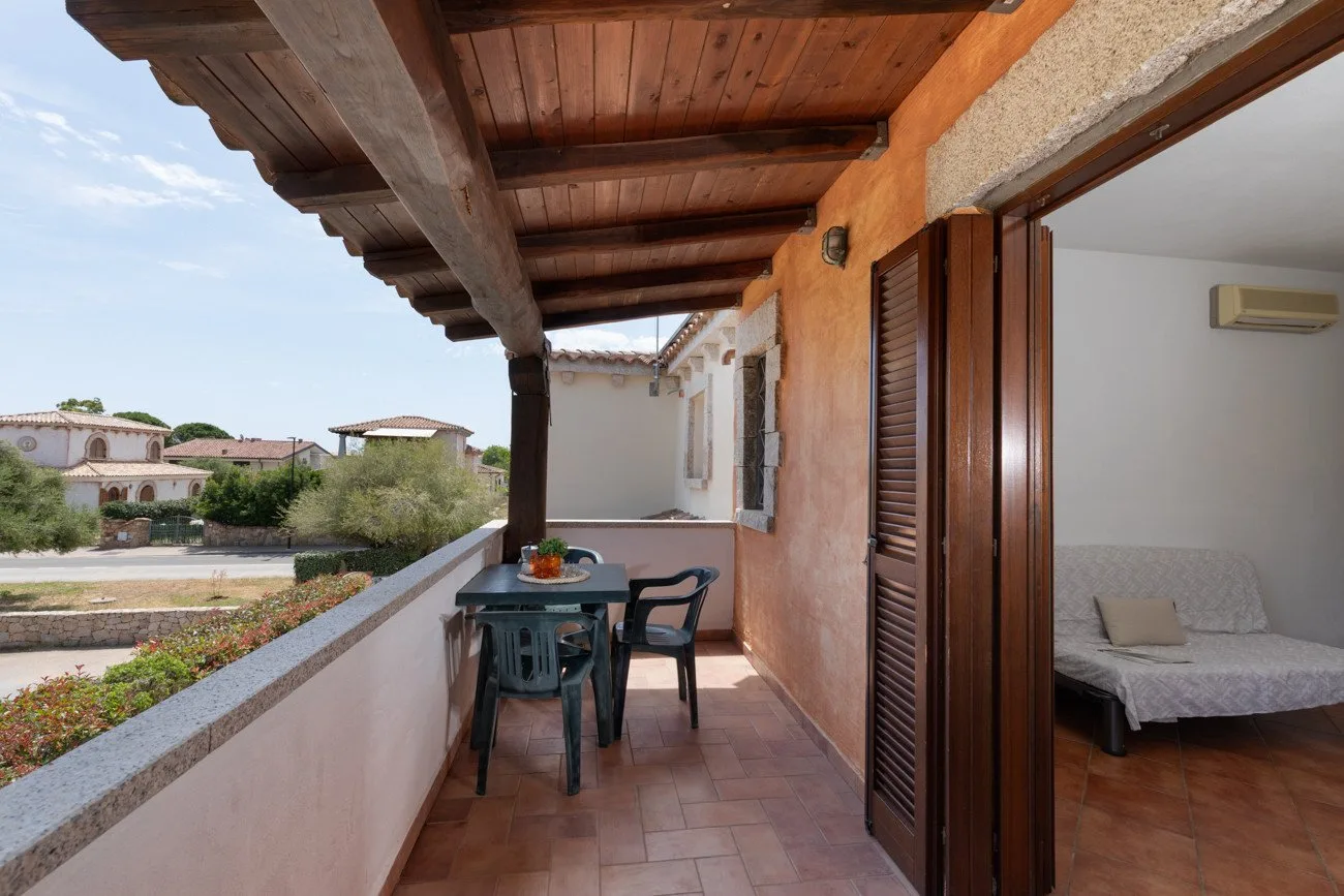 Una terraza cubierta tiene una mesa pequeña y sillas junto a una puerta abierta.