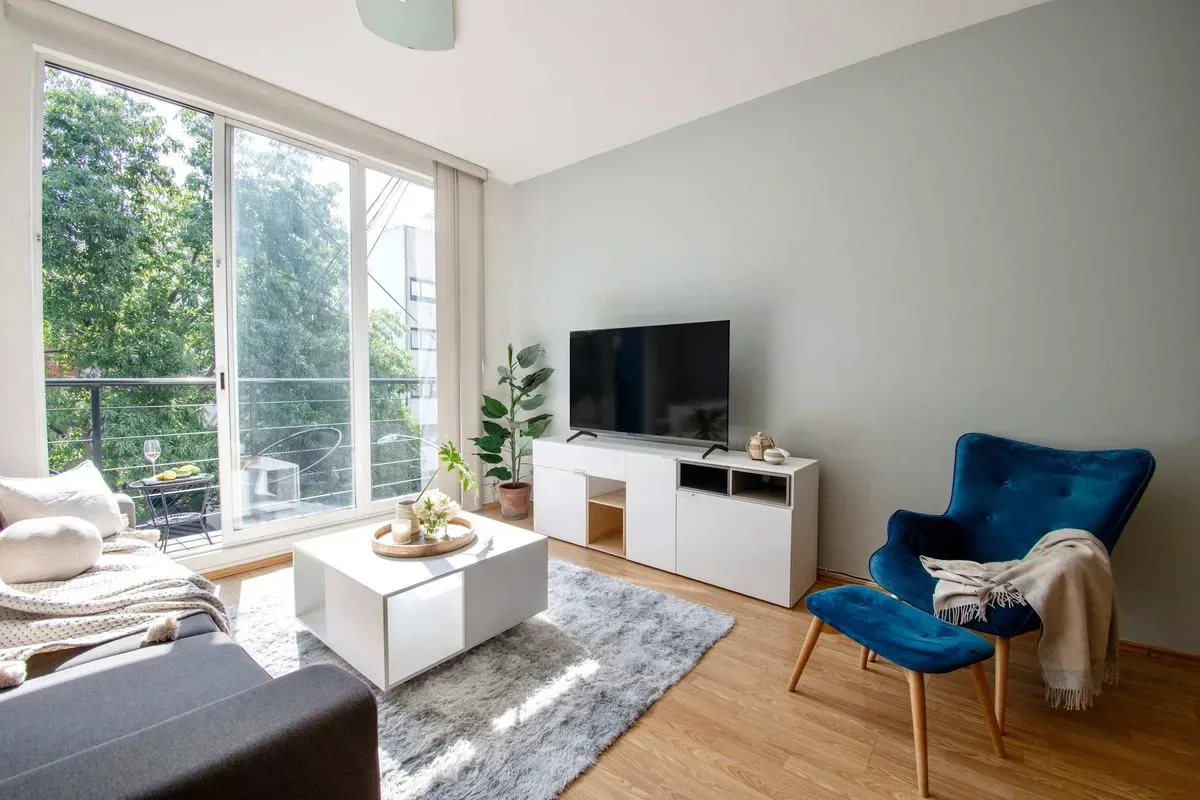 A bright living room with a television, seating, and large windows overlooking greenery.