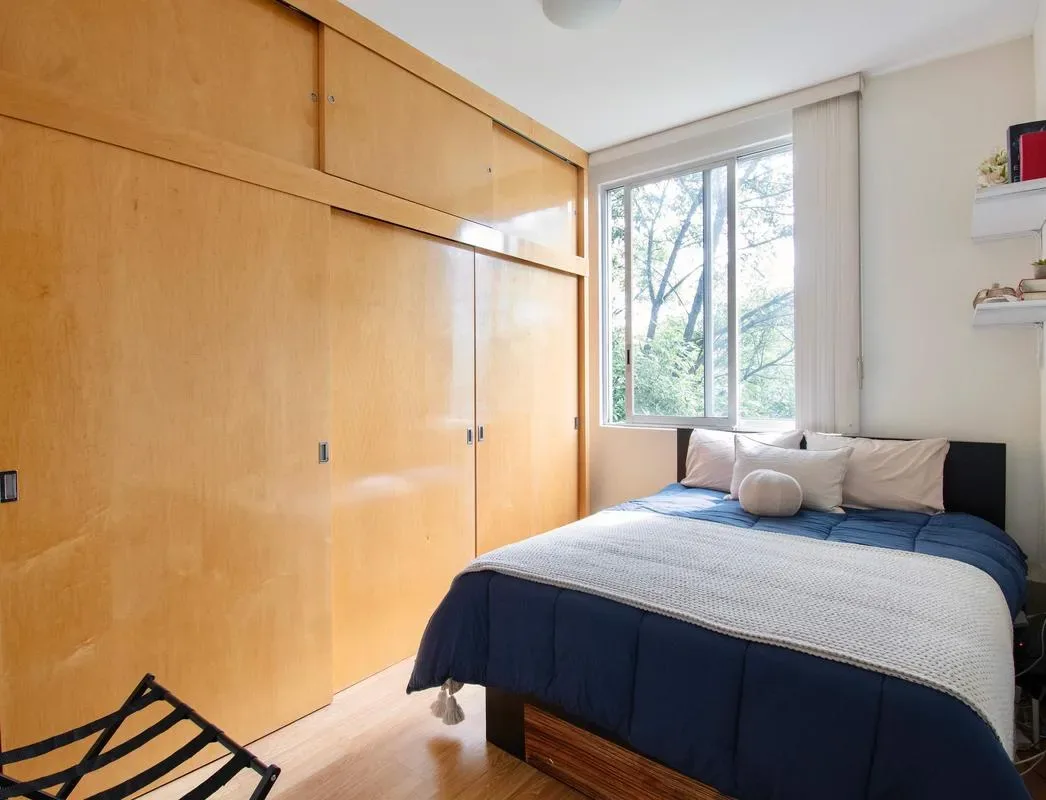 A bedroom with a bed beside a large window and built-in wooden storage.