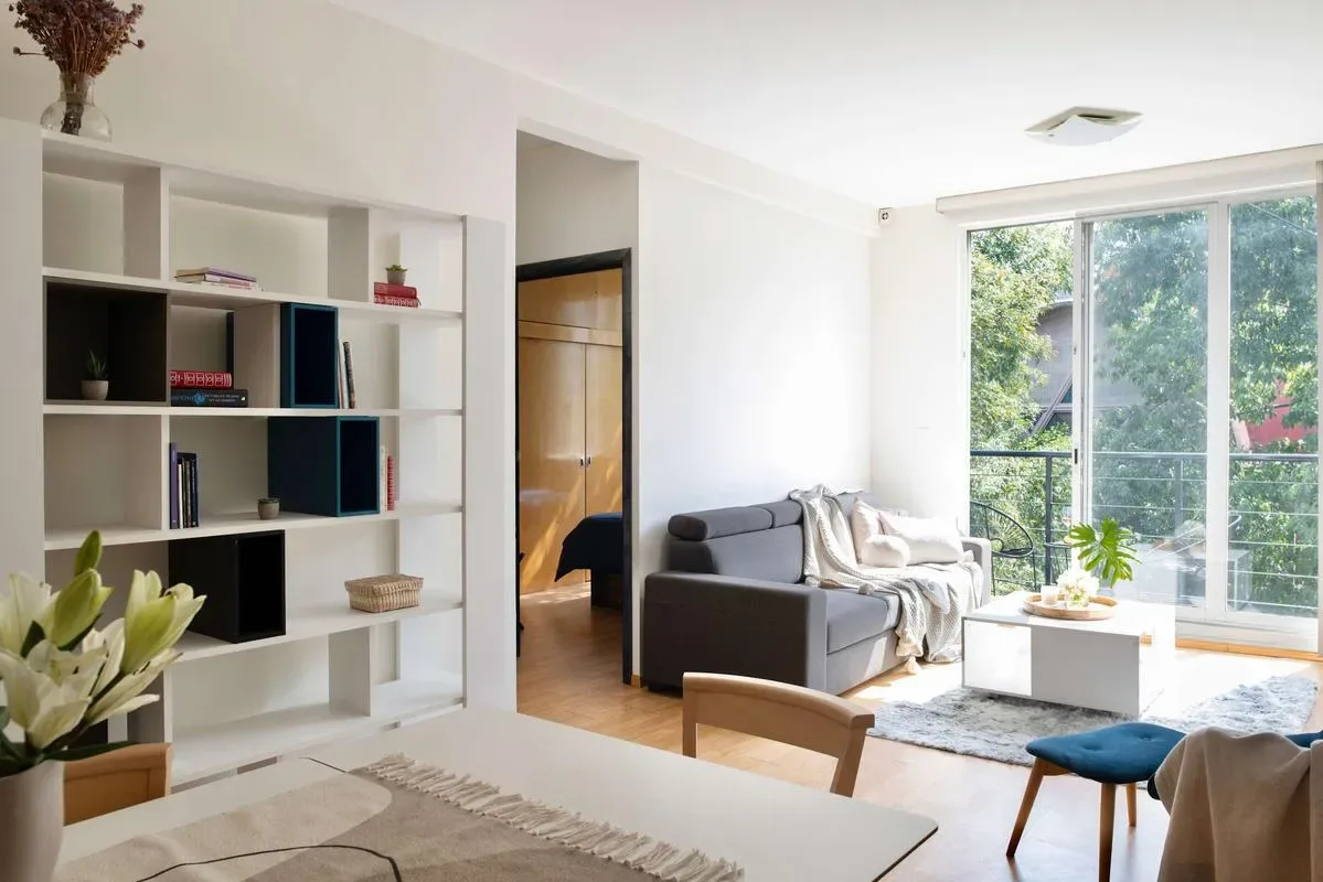 A bright living room with open shelving, seating, and large windows overlooking greenery.
