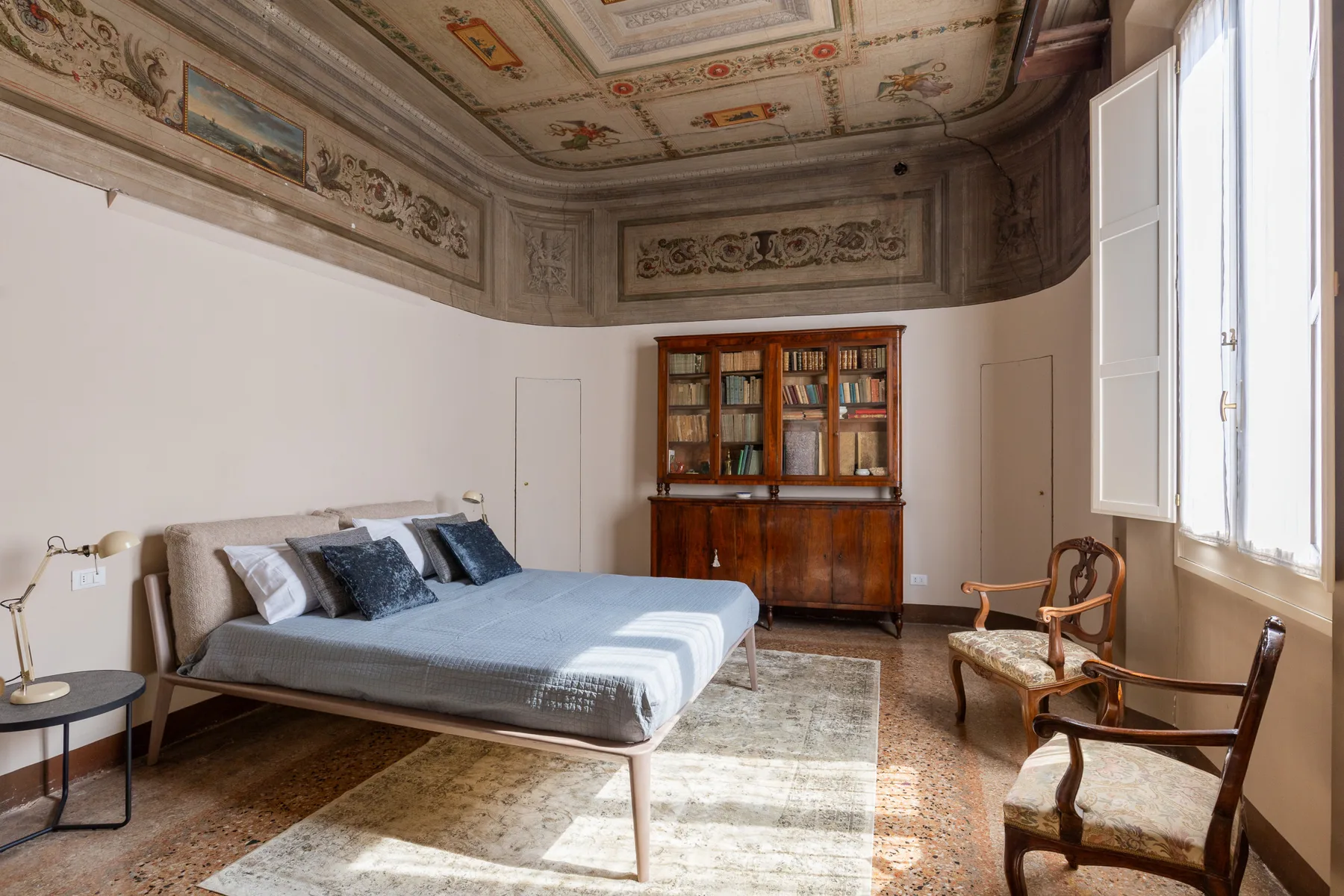 A bedroom with a bed, wooden cabinet, and chairs beneath a decorated ceiling.