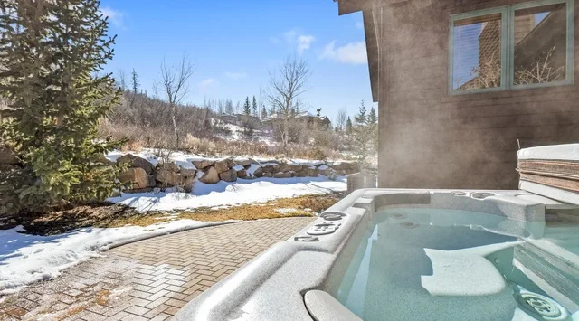 Un jacuzzi al aire libre está junto a un patio nevado y una casa.