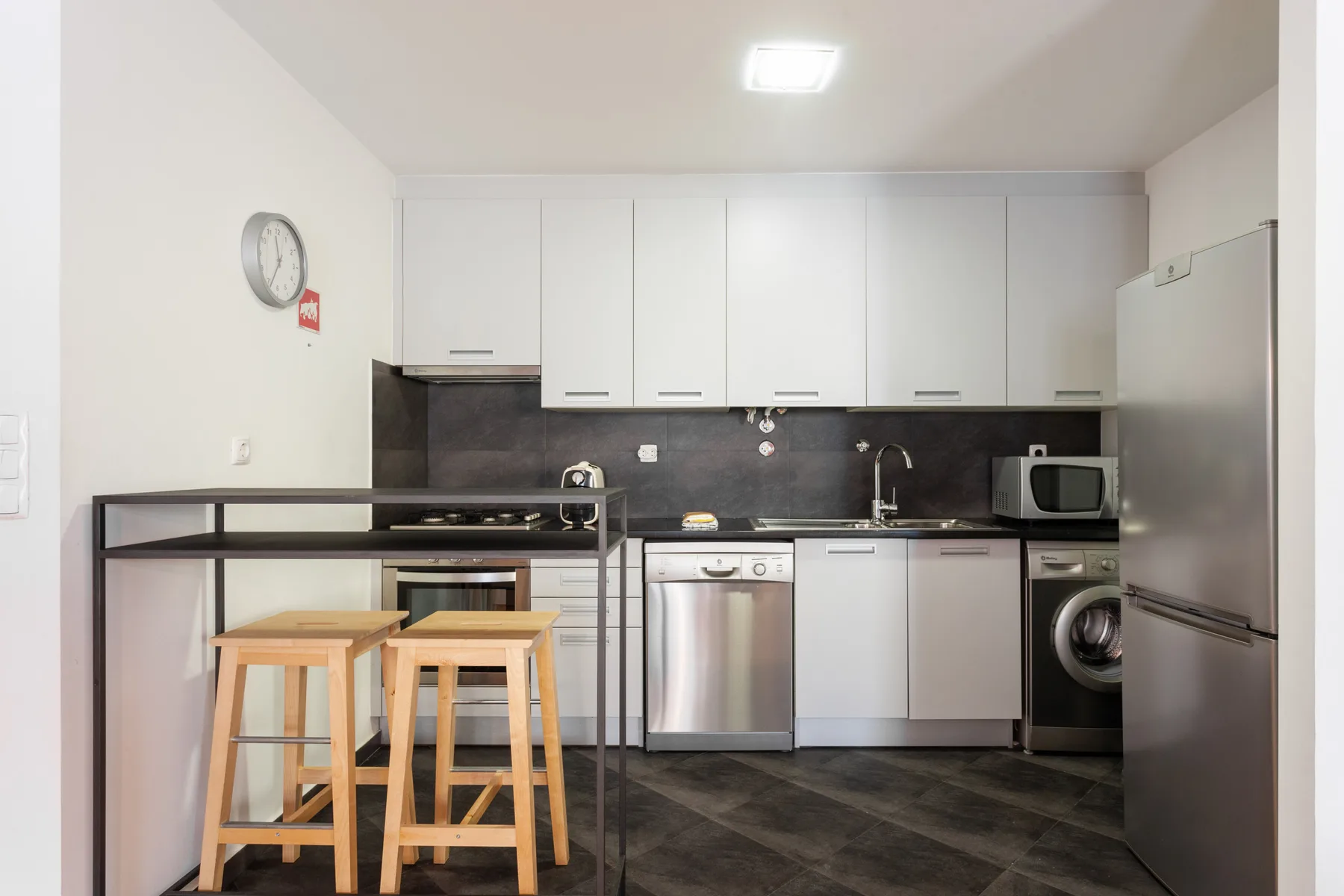 Una cocina compacta tiene gabinetes blancos, encimeras oscuras y una barra con taburetes.