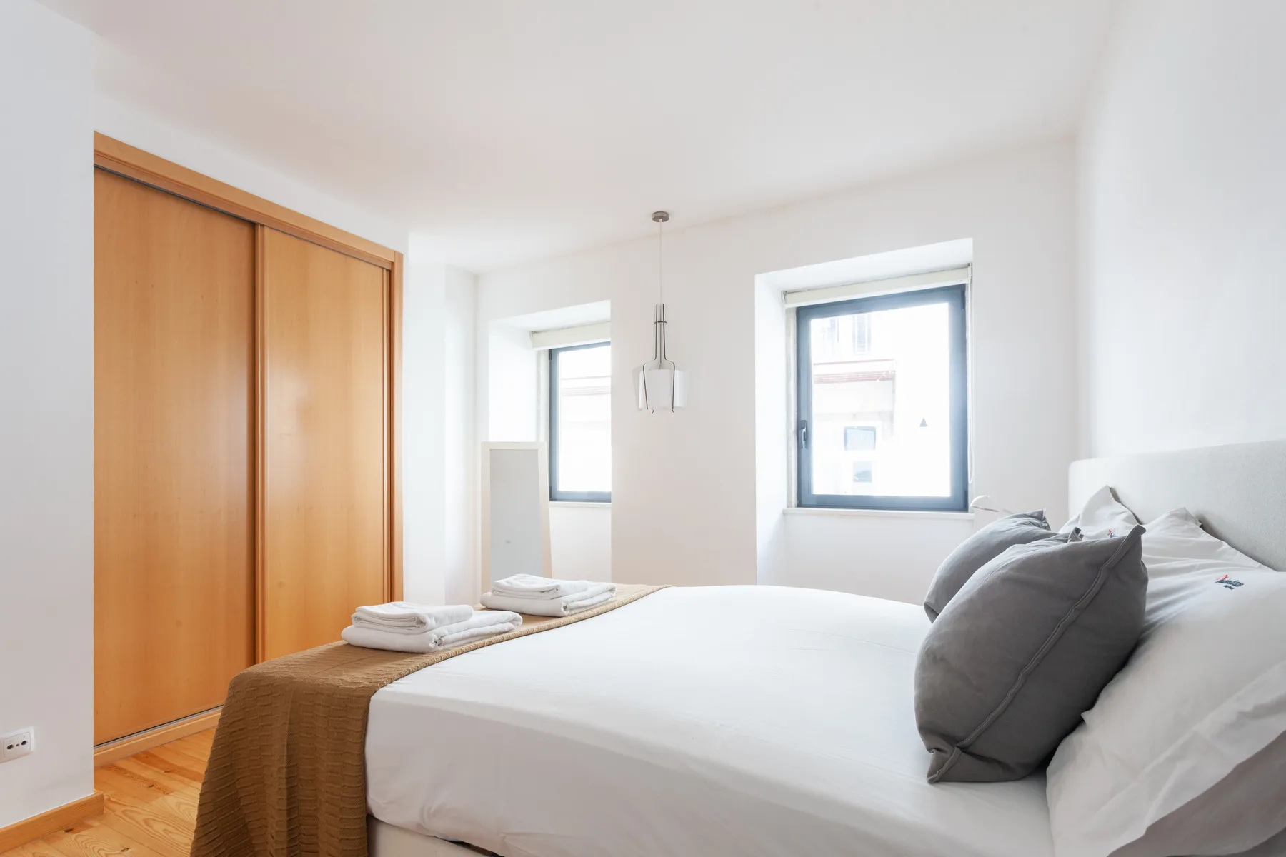 A bright bedroom with a bed, wooden closet, and two windows.