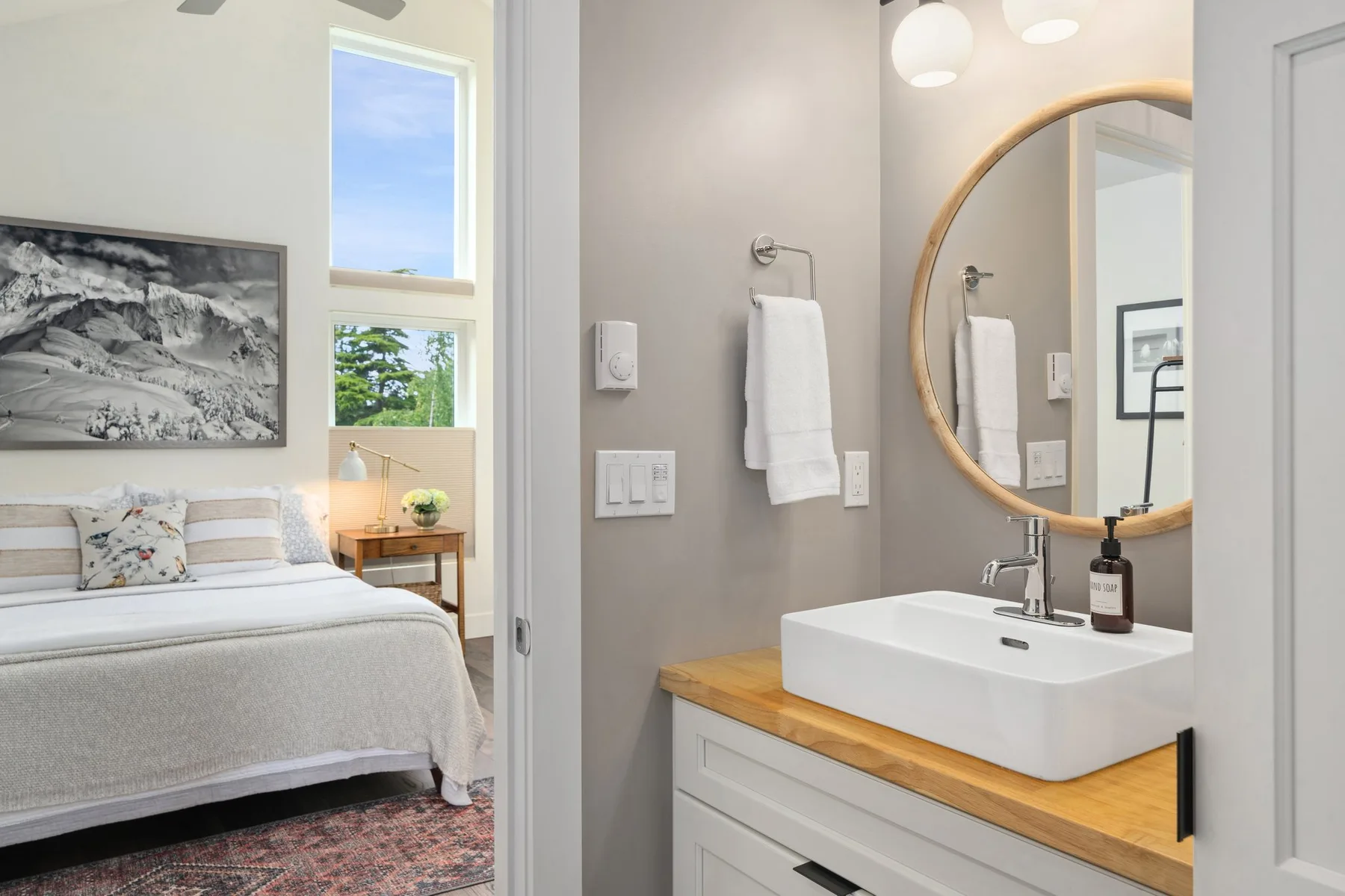 Un dormitorio con una cama bien arreglada, junto a un tocador de baño y un espejo redondo.