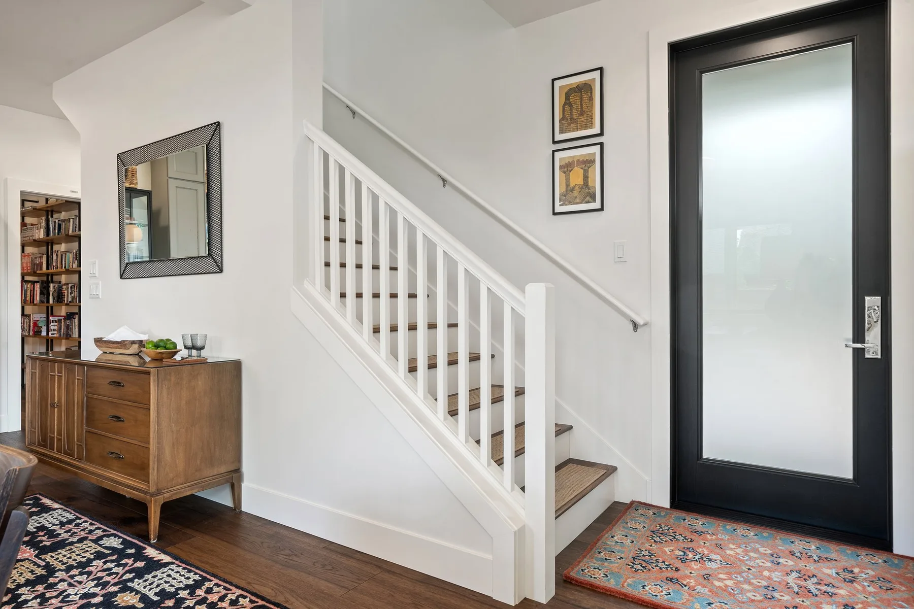 Una entrada luminosa con una escalera, un mueble de madera y una puerta con panel de vidrio.