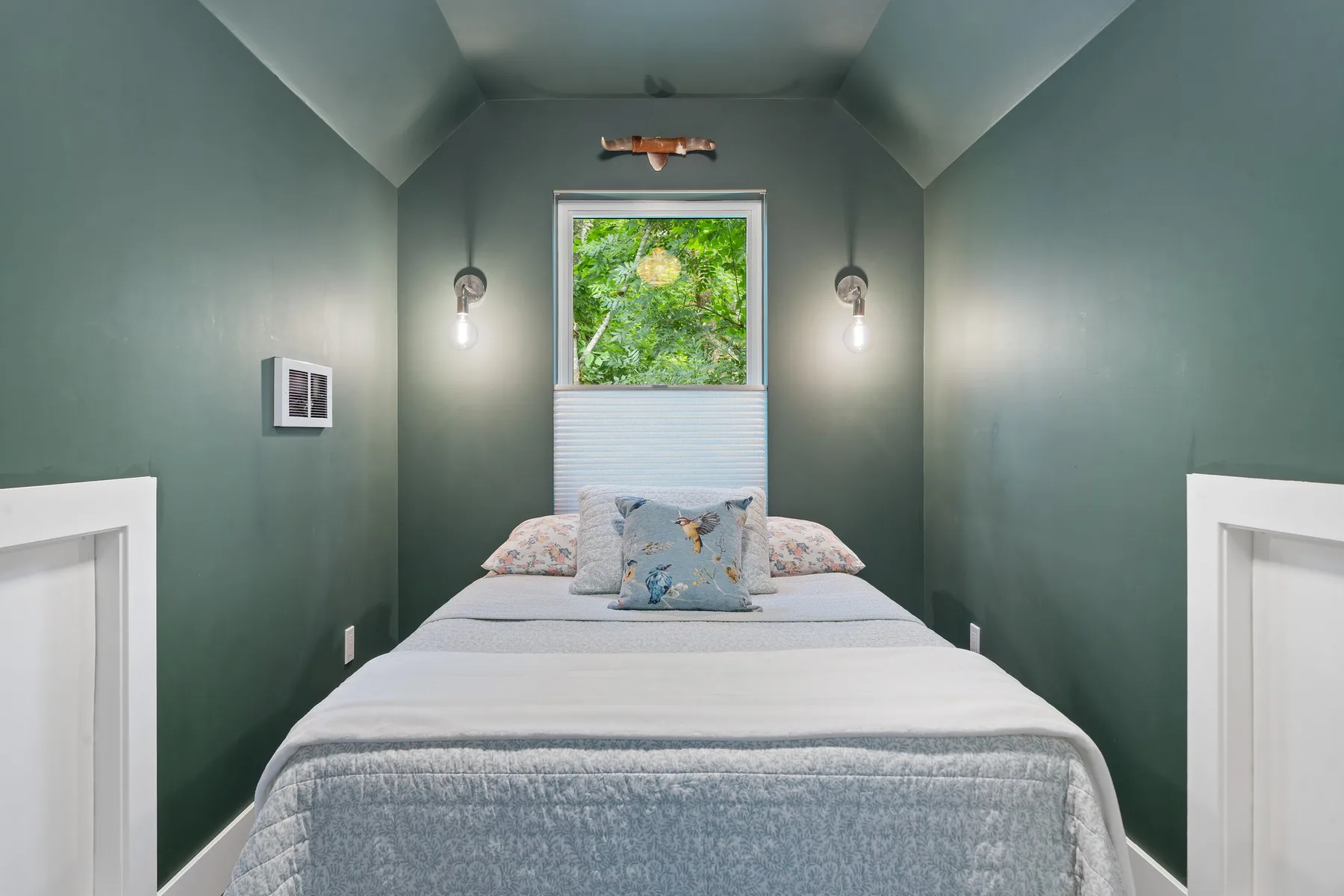 Un dormitorio compacto con una cama bajo una ventana que da a un follaje frondoso.
