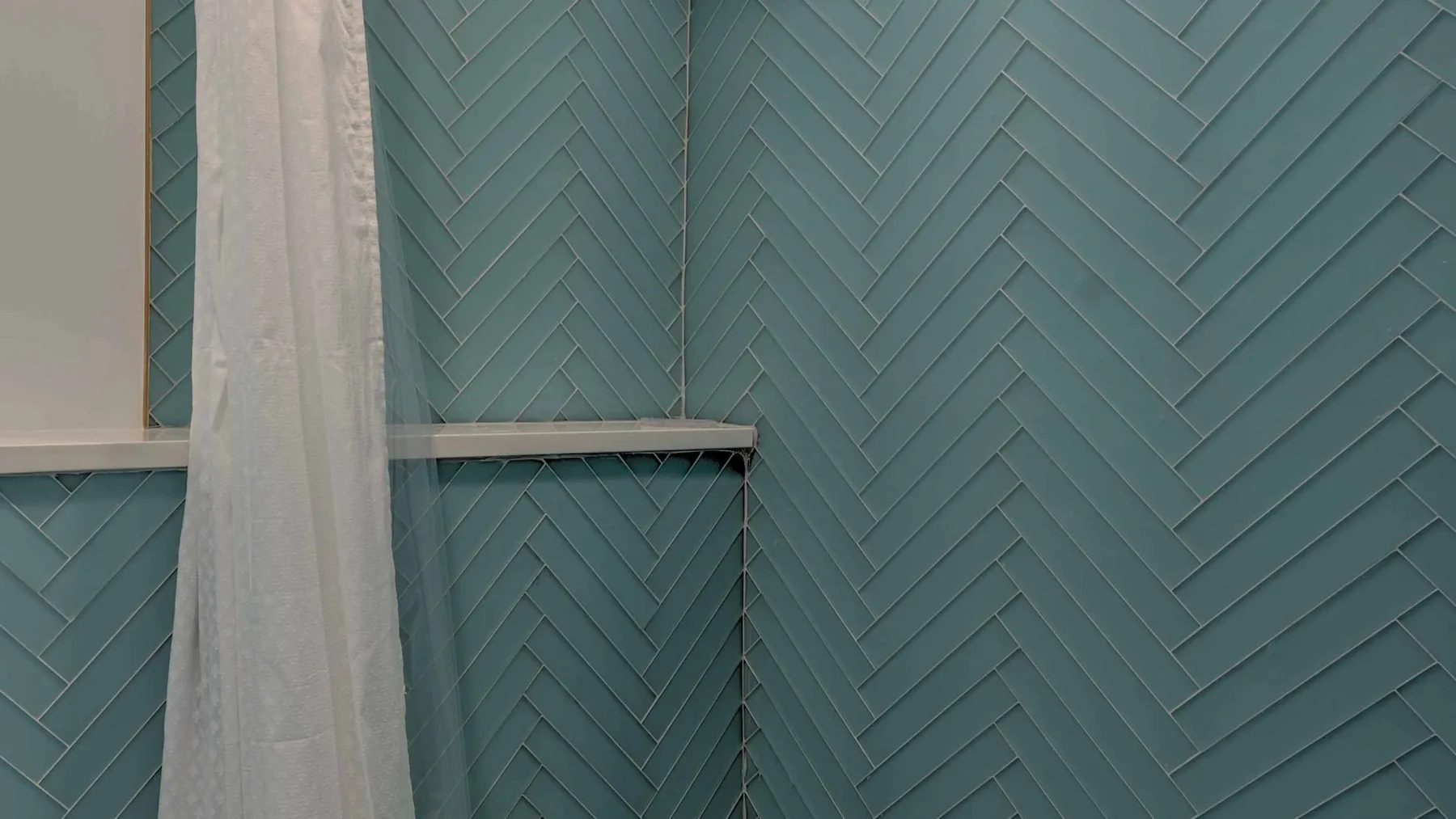 Una zona de ducha con azulejos verde azulado en espiga y una cortina clara.
