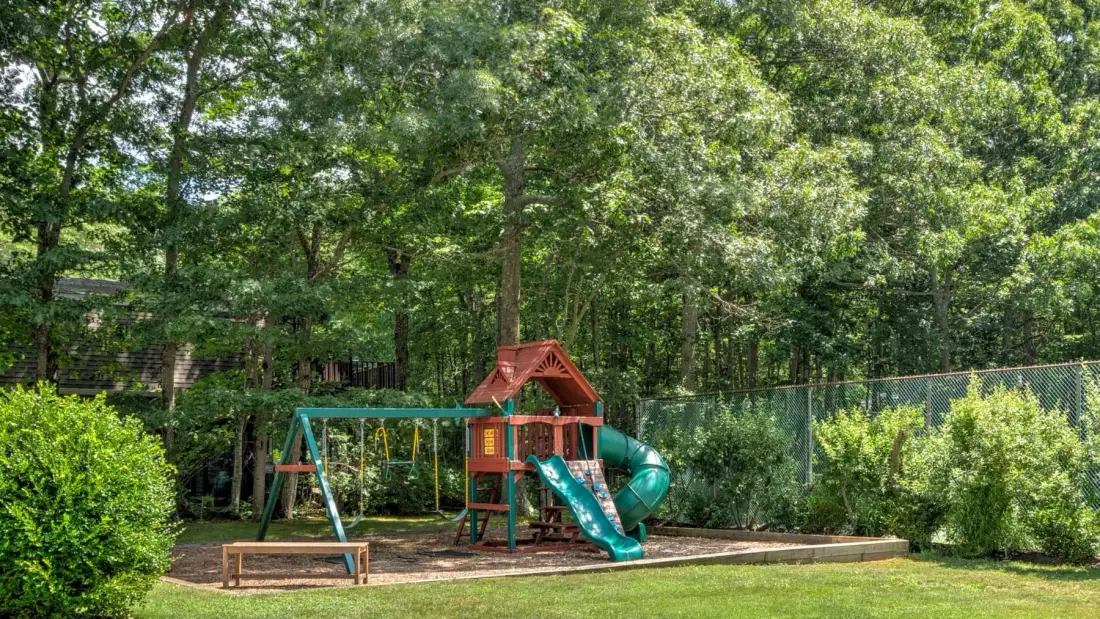 Un área de juegos al aire libre entre árboles cuenta con un tobogán, columpios y una estructura para trepar.