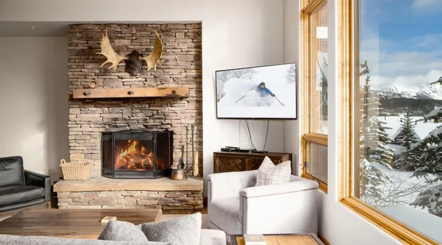 Una sala de estar con chimenea de piedra y grandes ventanas con vistas a un paisaje nevado.