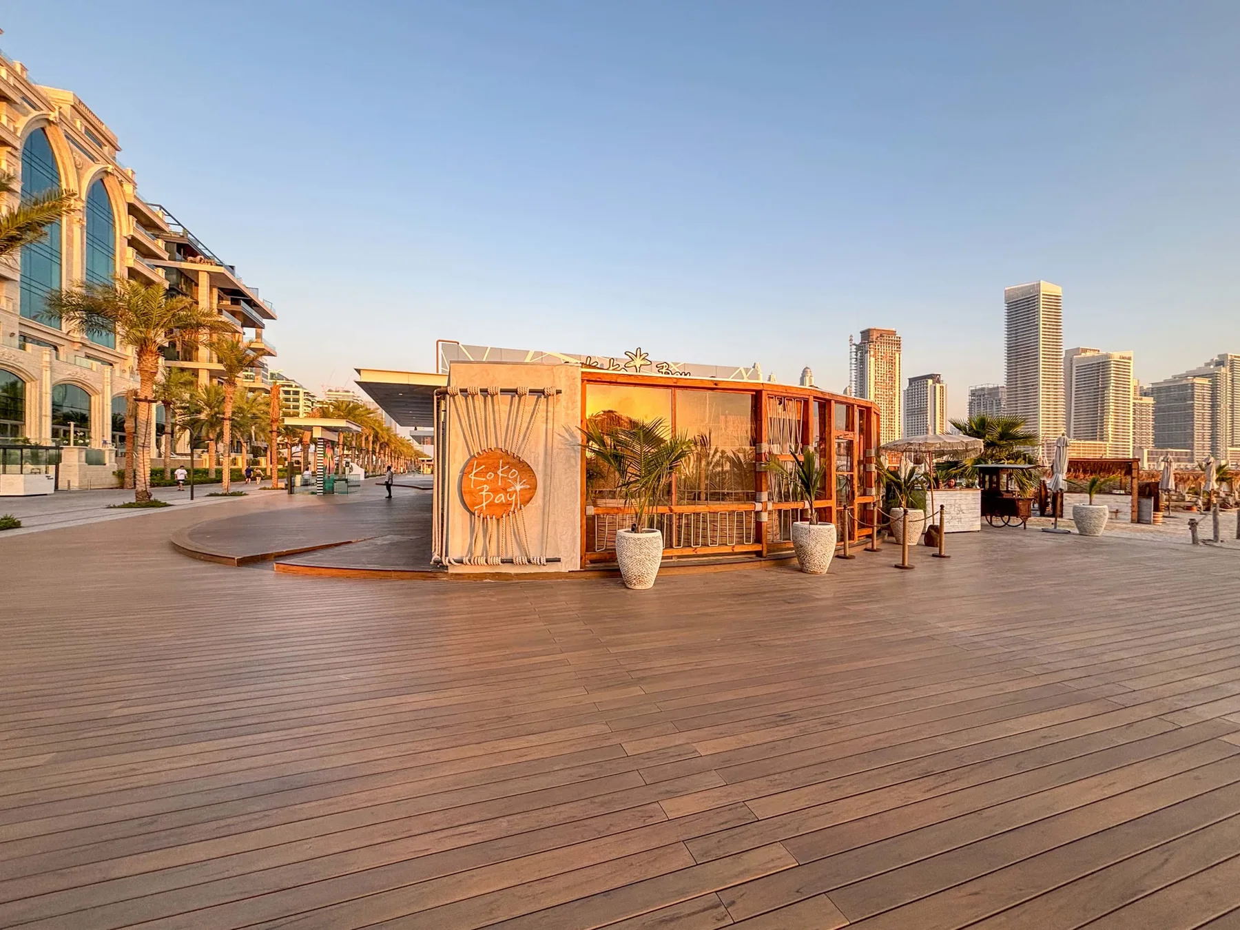 Una terraza de madera junto al agua, con un pequeño comedor cerrado y edificios al fondo.
