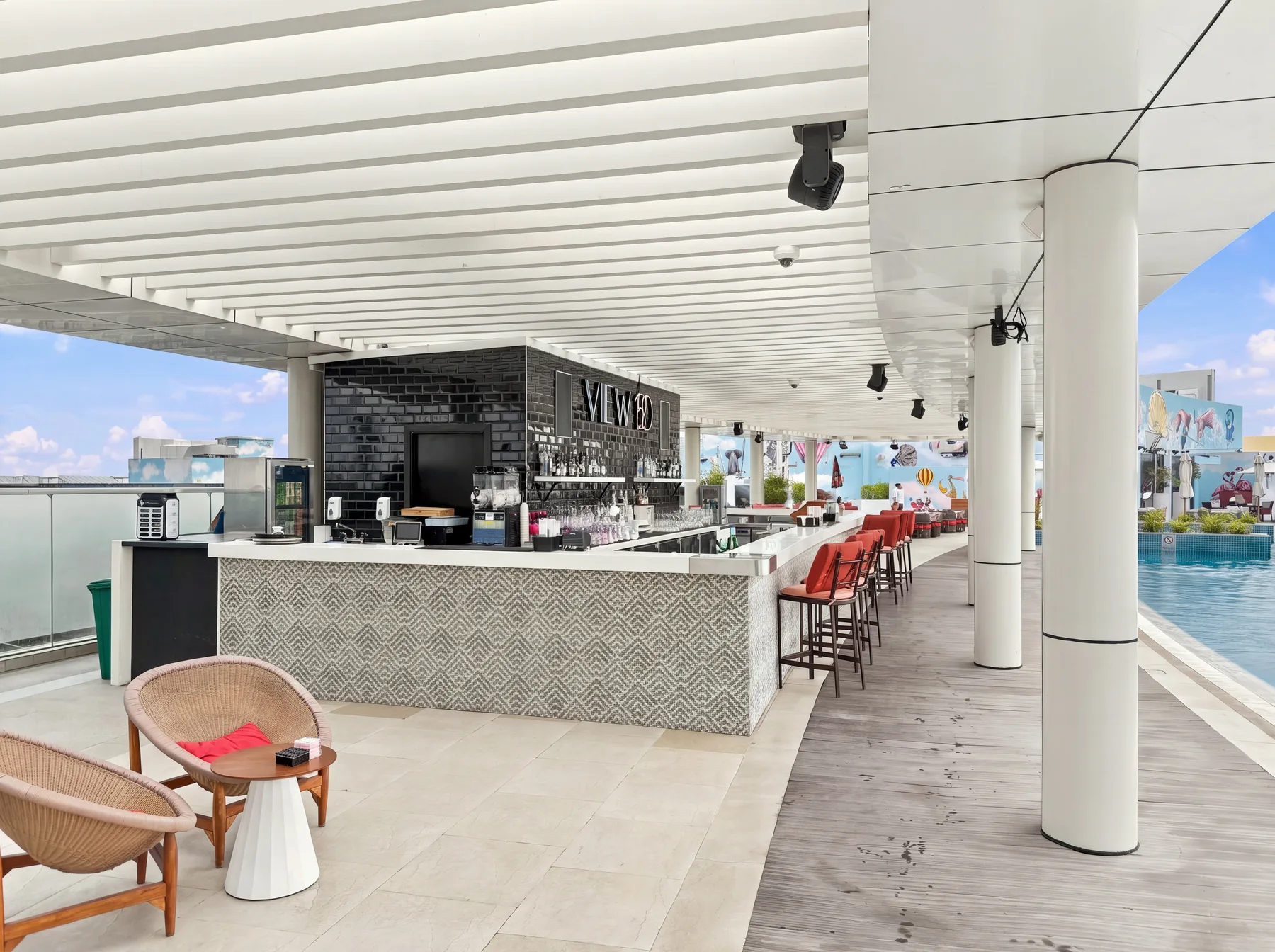 Un bar cubierto junto a la piscina tiene una barra estampada, taburetes rojos y asientos cercanos.