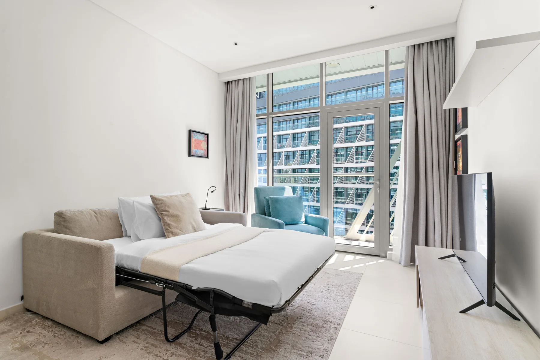 Un dormitorio luminoso con sofá cama junto a grandes ventanas y un televisor.