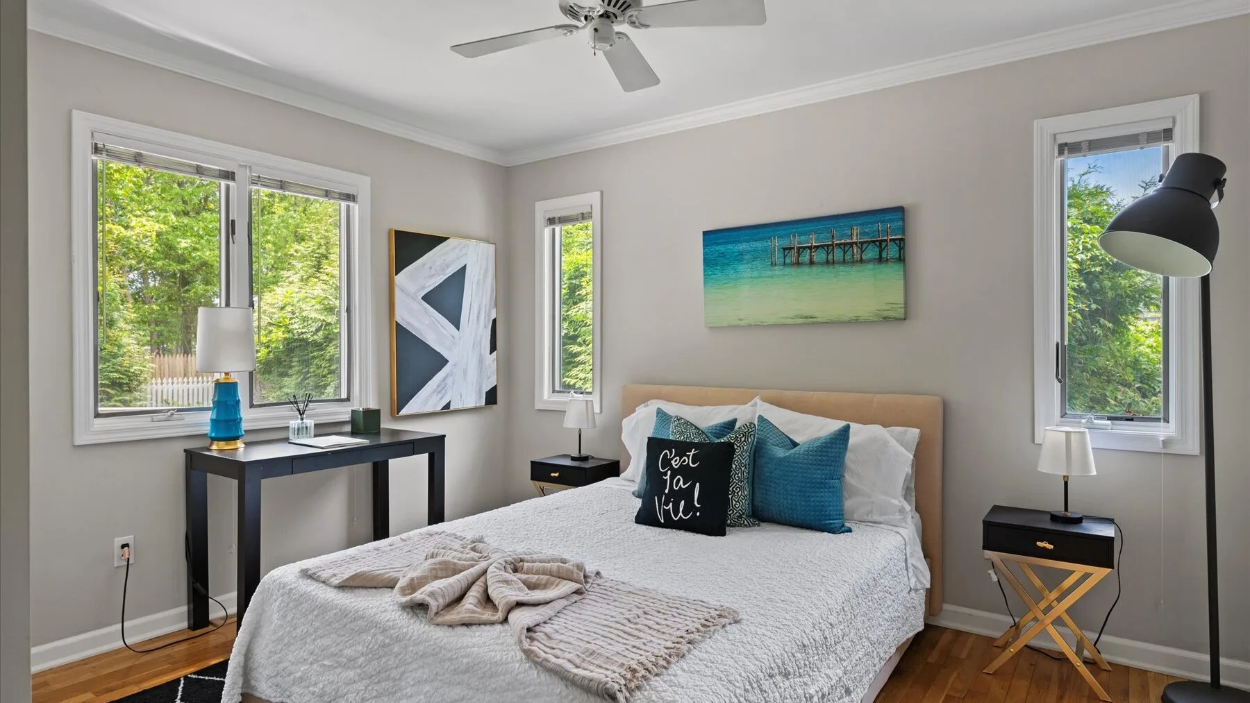A bright bedroom with a bed, blue accent pillows, and windows overlooking greenery.