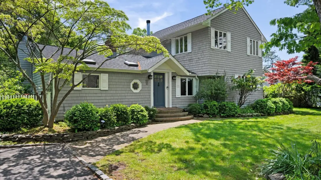 A gray-sided house stands amid a green lawn, trees, and garden shrubs.