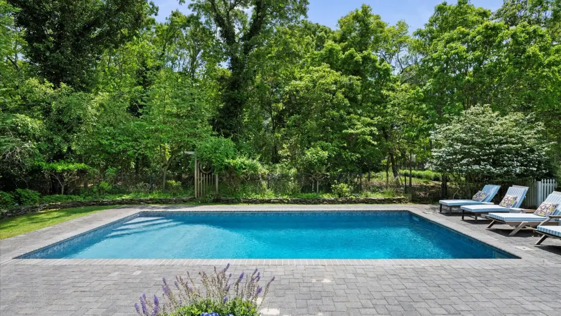 An outdoor swimming pool sits beside a paved patio and leafy trees.