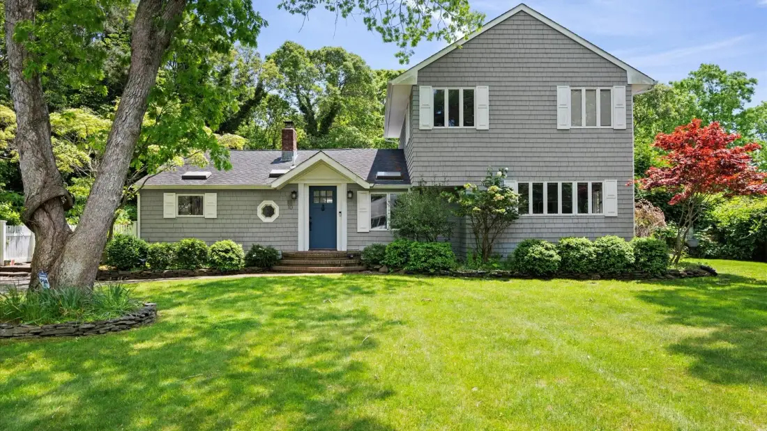 A two-story house stands behind a green lawn, with trees and shrubs around it.