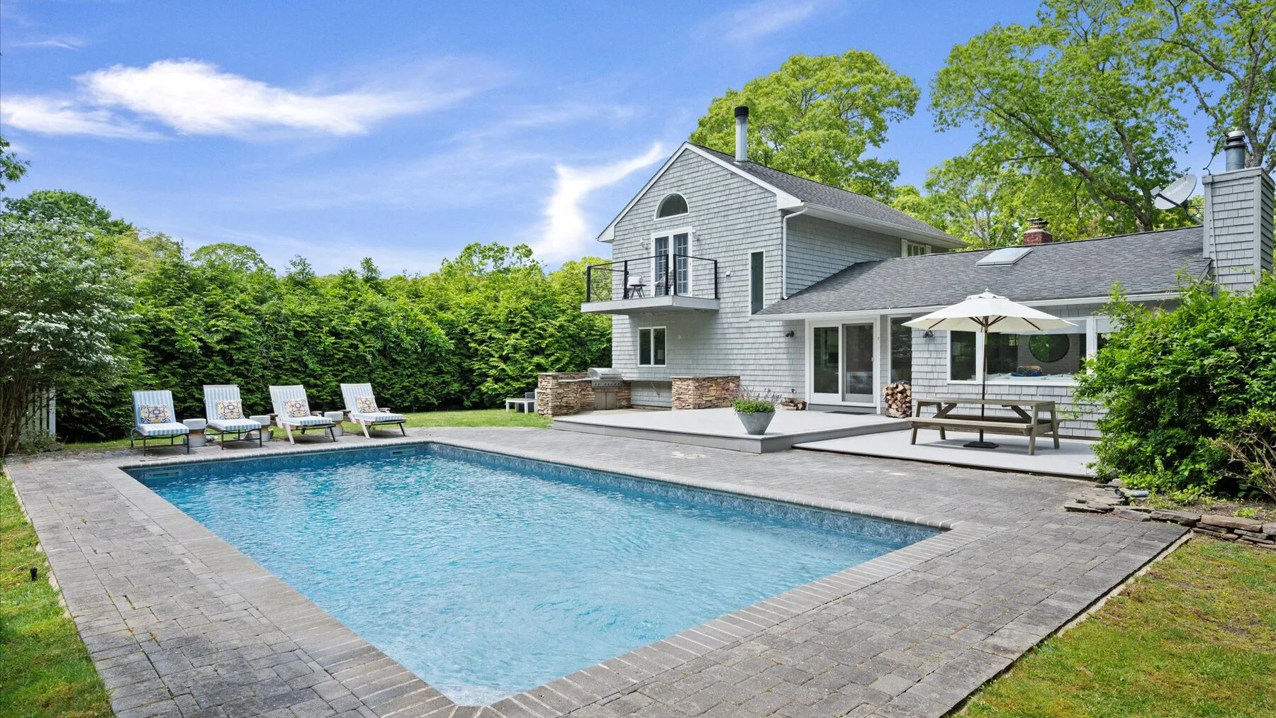 An outdoor swimming pool sits beside a paved patio and a large house.