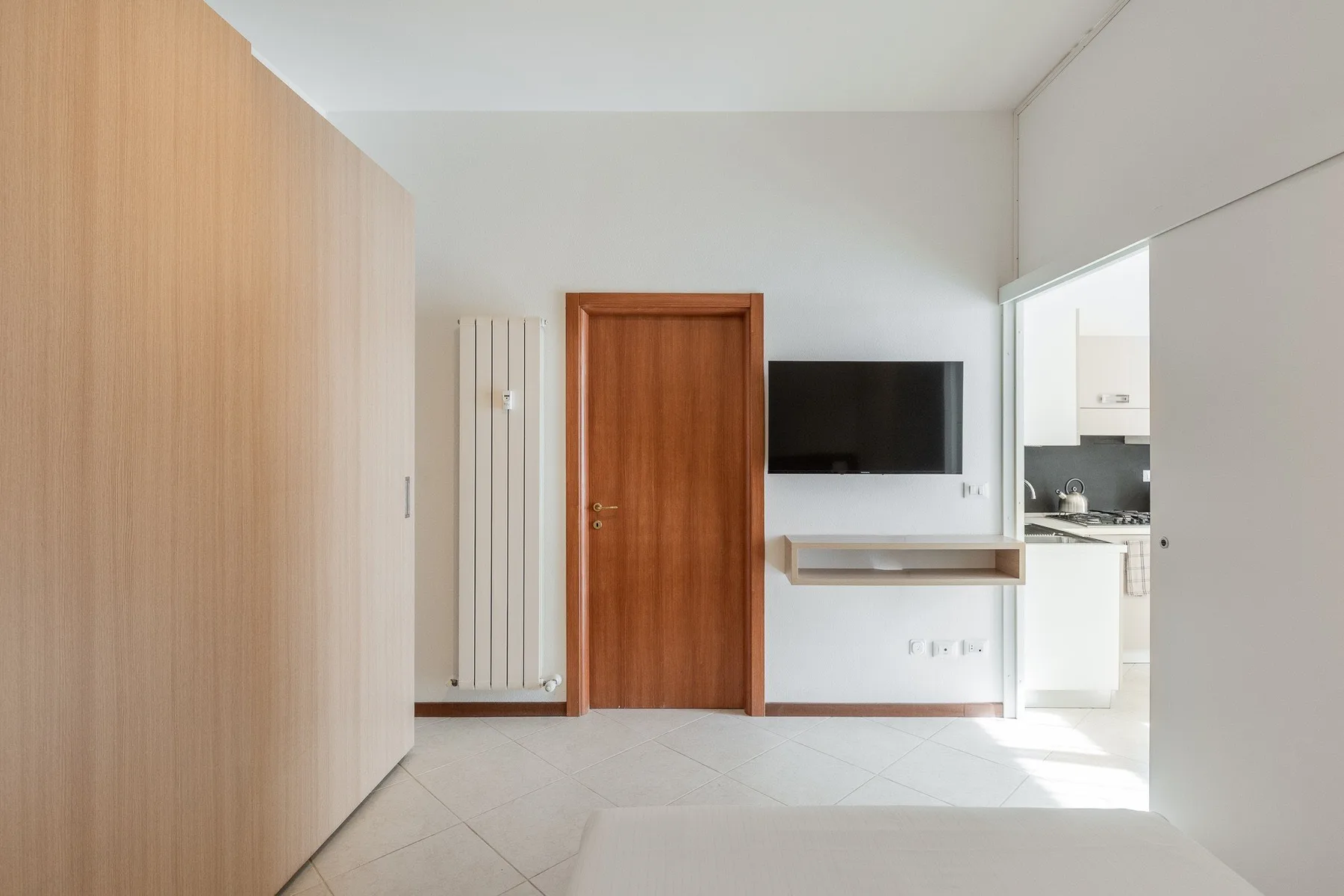 A bright room with a wooden door, wall-mounted television, and an open kitchen doorway.