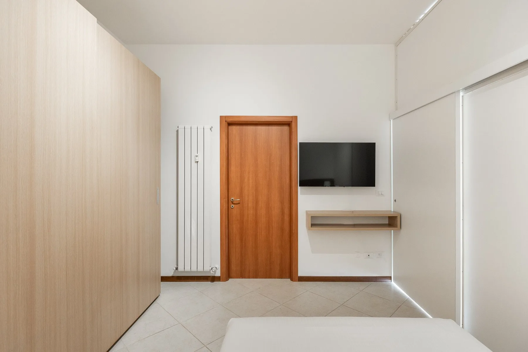 A bedroom with a wooden door, wall-mounted television, and a bed in the foreground.