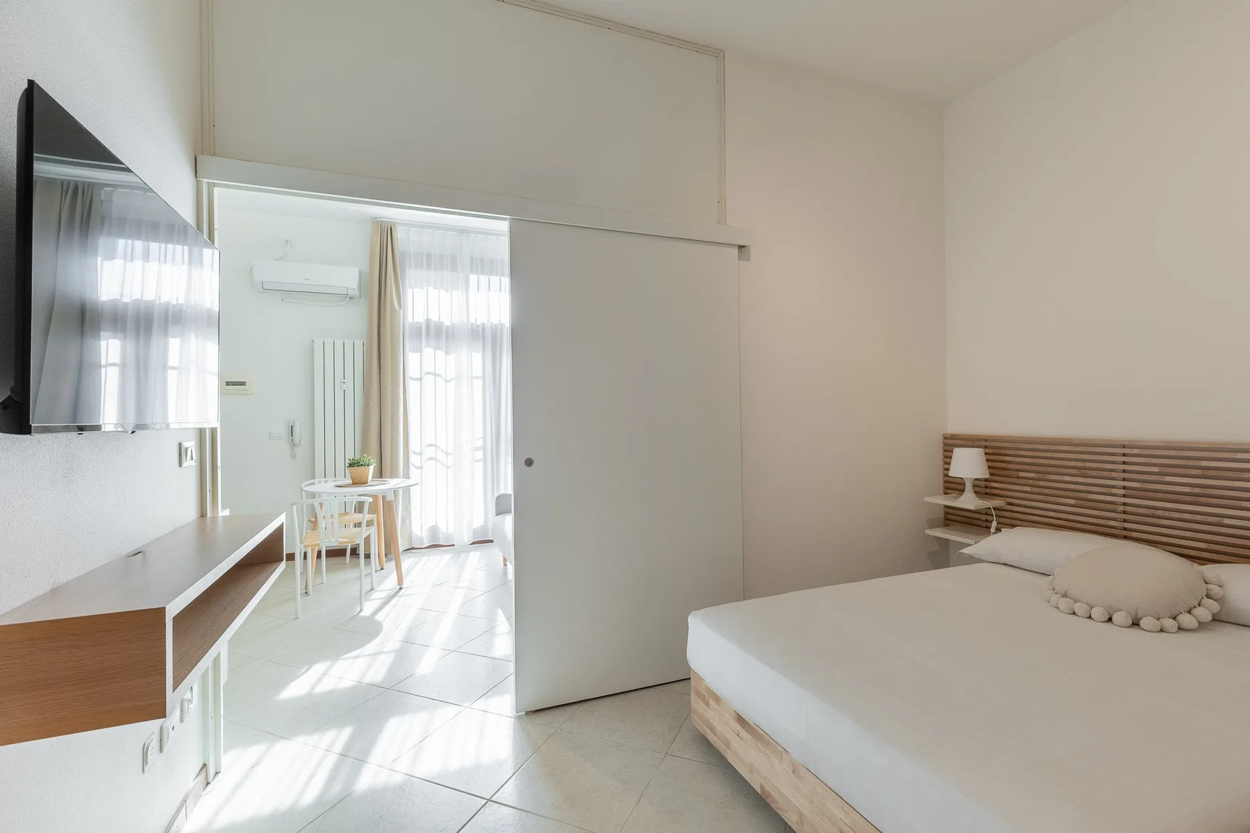 A bright bedroom with a bed, wall-mounted television, and sliding door to a dining area.