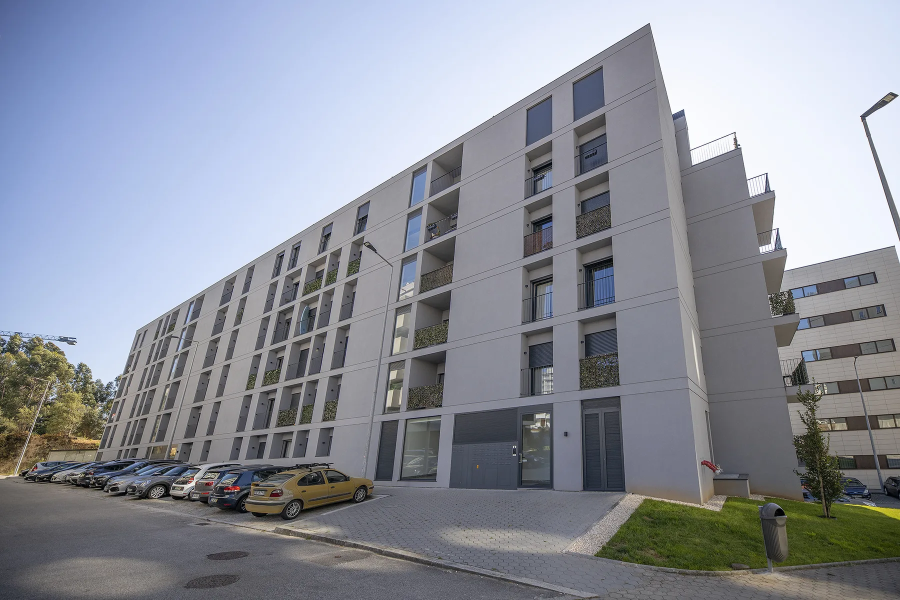 A modern apartment building faces a paved parking area with parked cars.