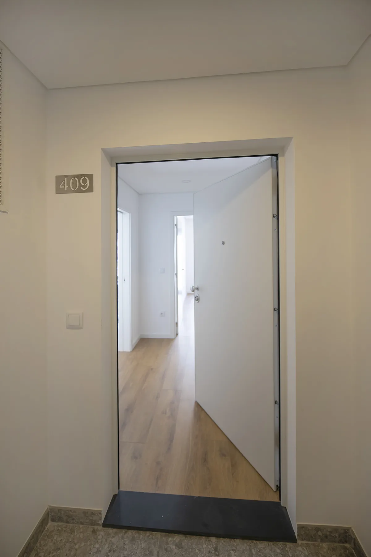 Una puerta da a un pasillo luminoso con suelo de madera clara.