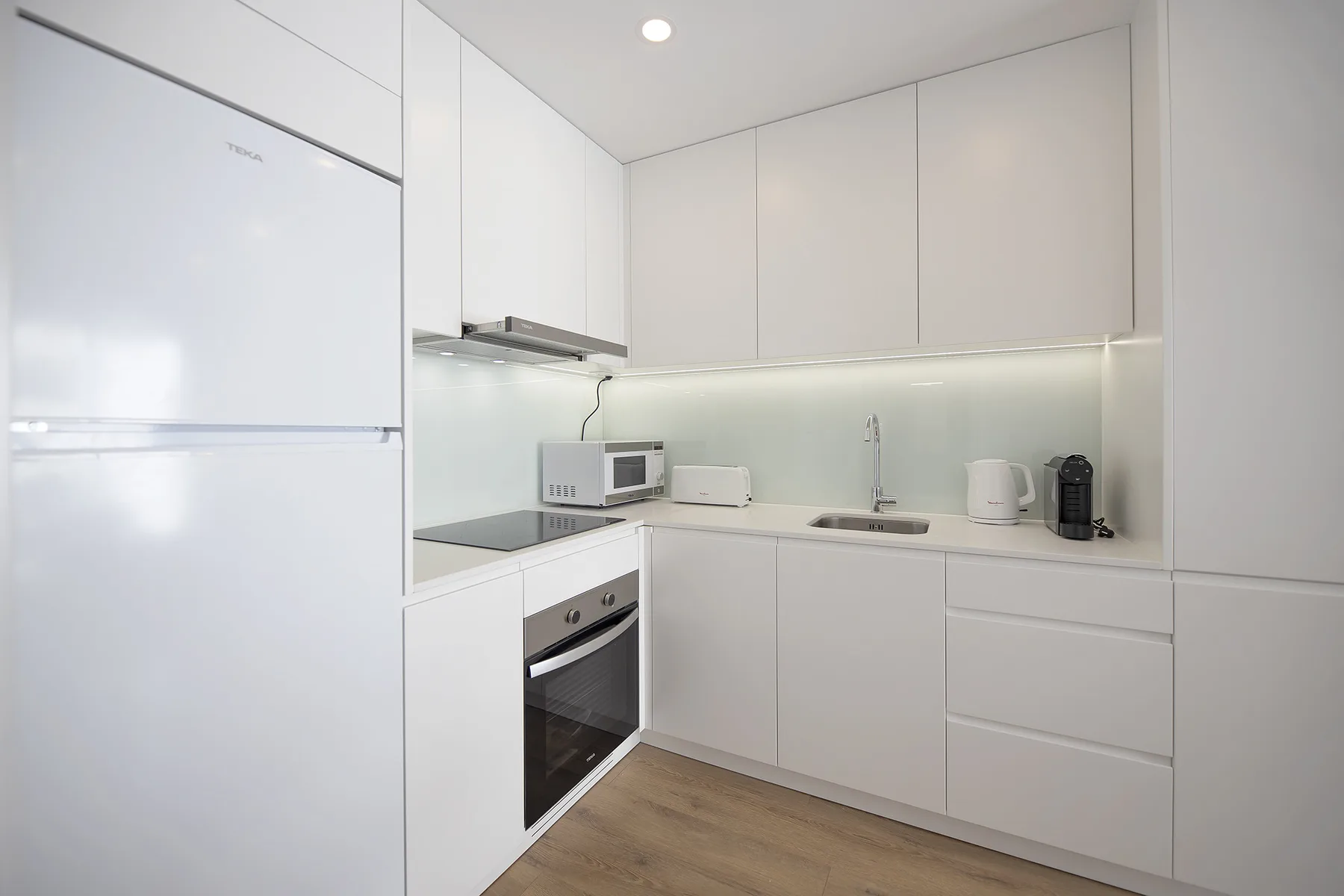 Una cocina blanca y luminosa con refrigerador, horno y pequeños electrodomésticos sobre la encimera.