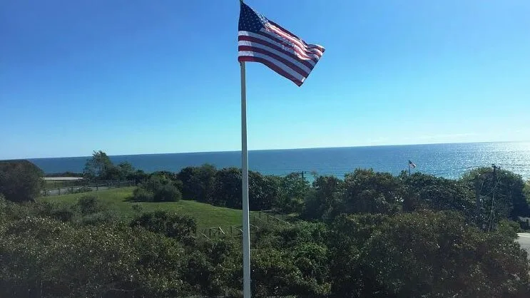 Una bandera de Estados Unidos ondea sobre la vegetación costera, con el océano al fondo.