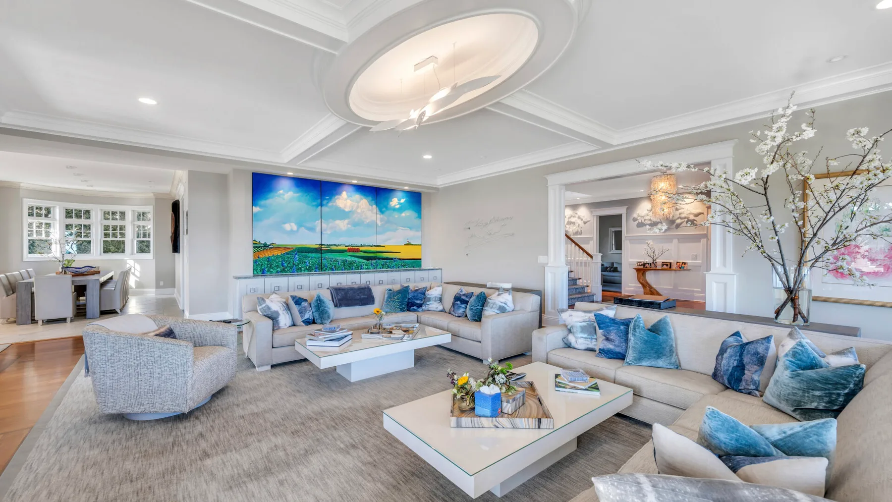 A bright living room with pale seating, low tables, and colorful wall art.
