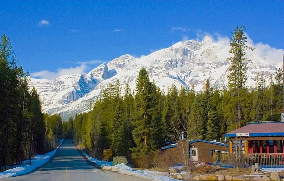 Lodge beside a paved road, evergreen forest and snow-capped mountains.