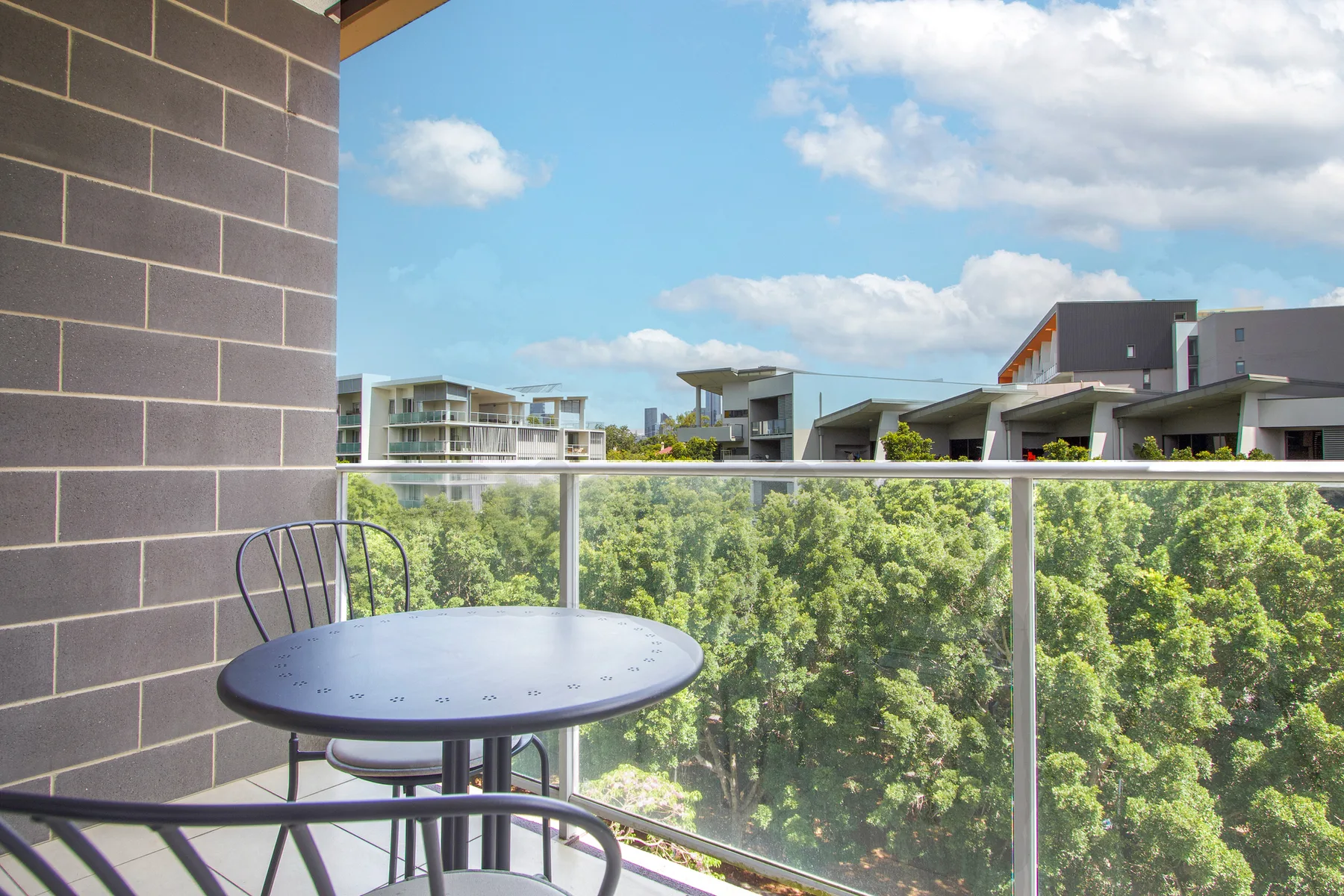 A balcony with a round table and chairs overlooks leafy trees and nearby buildings.