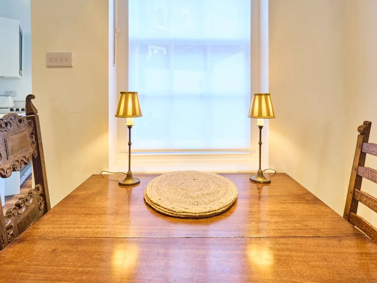 A wooden dining table sits beneath a bright window, with two lamps and a woven centerpiece.