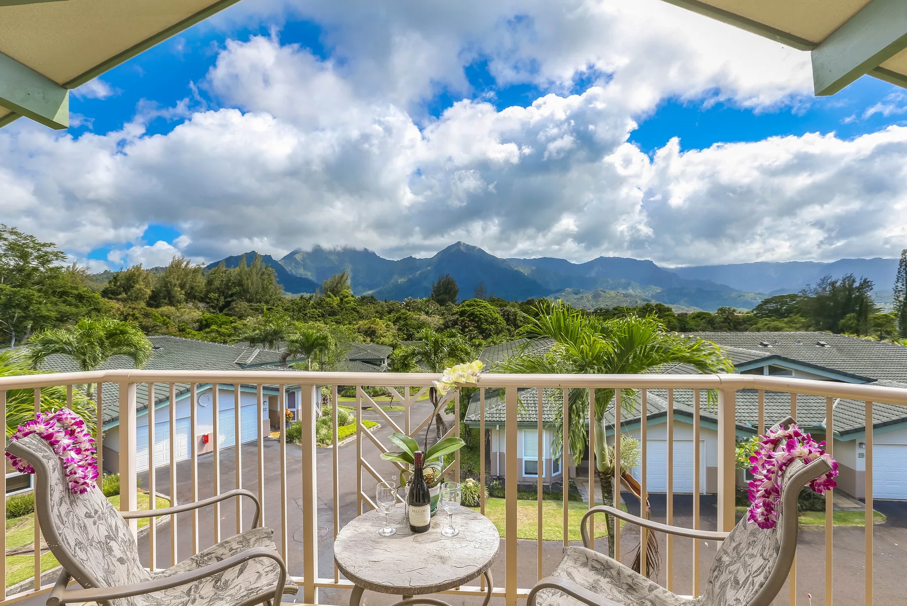 A balcony overlooks rooftops, greenery, and distant mountains beneath a cloudy sky.