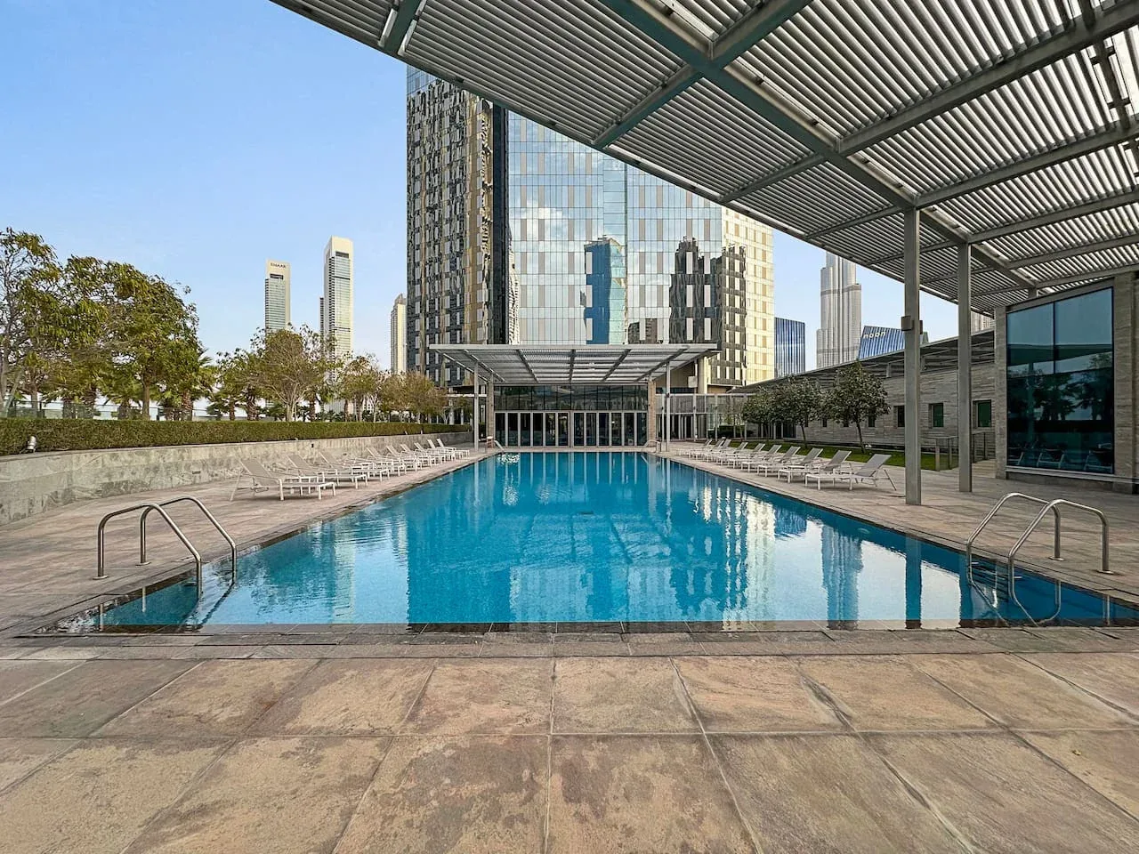 Una piscina al aire libre está bajo una cubierta de listones, con edificios urbanos al fondo.