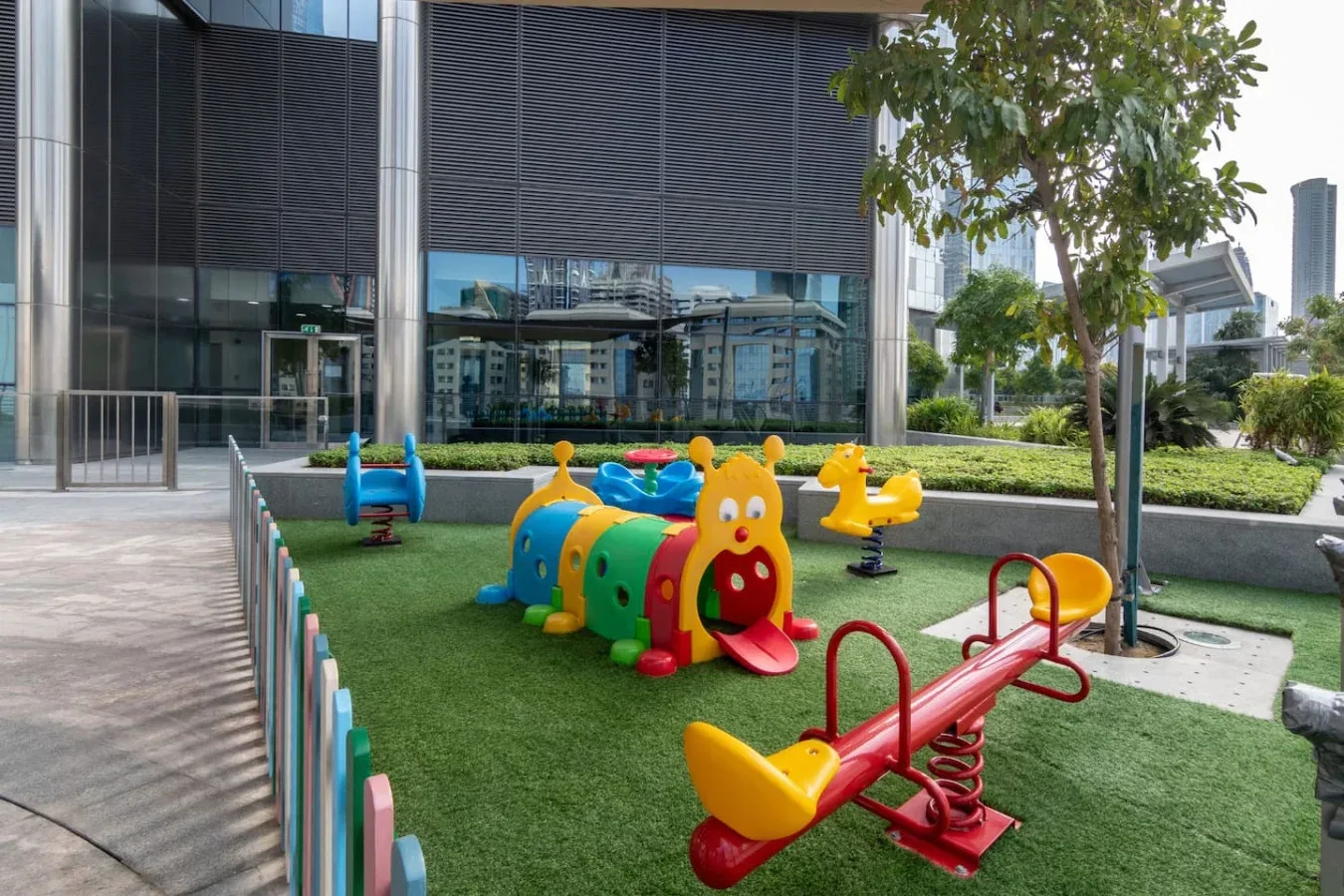 Un patio de juegos cercado con equipos coloridos está junto a un edificio con fachada de vidrio.