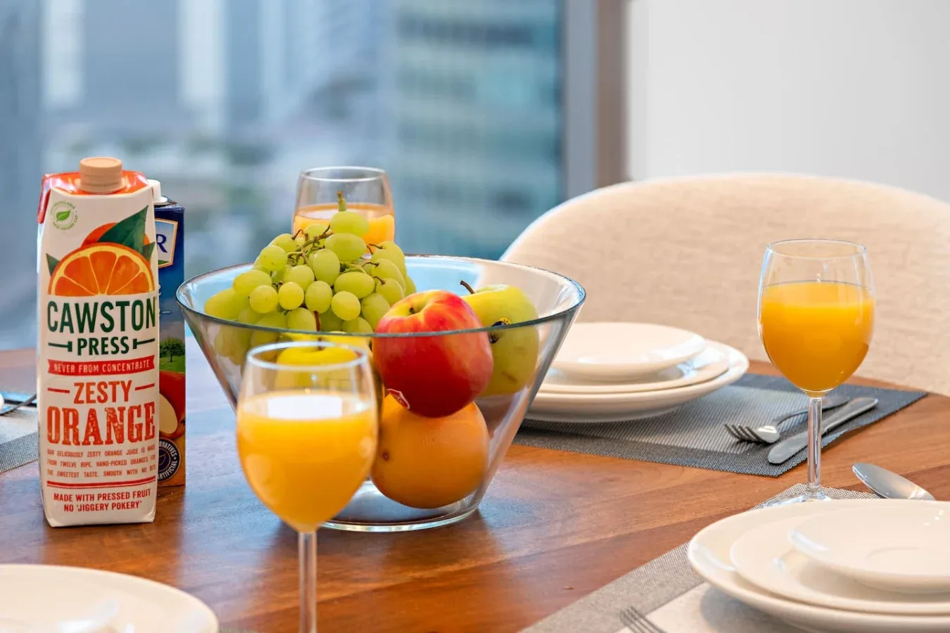 Una mesa de comedor está puesta con platos, vasos de zumo de naranja y un cuenco de fruta.