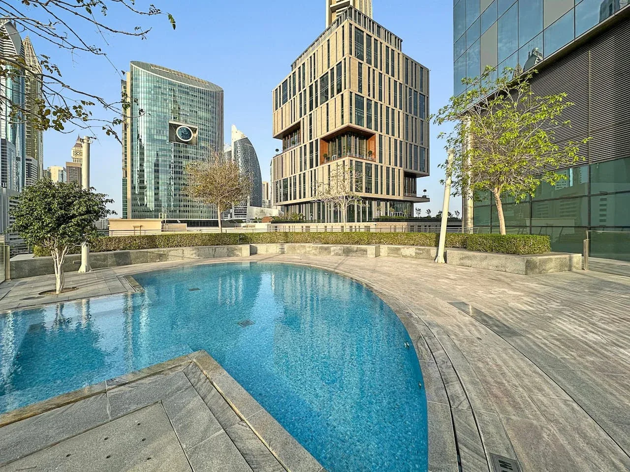 Una piscina al aire libre junto a una terraza pavimentada, con edificios modernos al fondo.